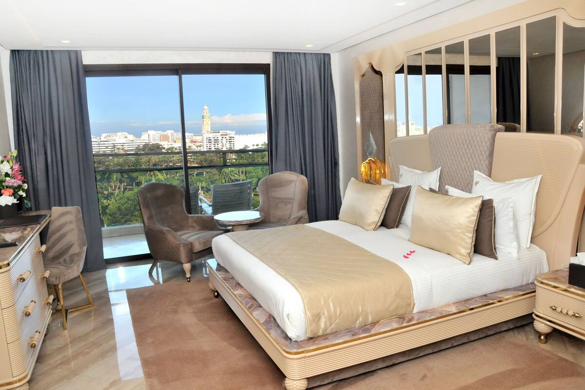 Ein geräumiges Schlafzimmer mit einem großen Bett, einer Sitzecke mit einem Sessel und einem kleinen Tisch sowie einem Blick auf die Stadt durch große Fenster.