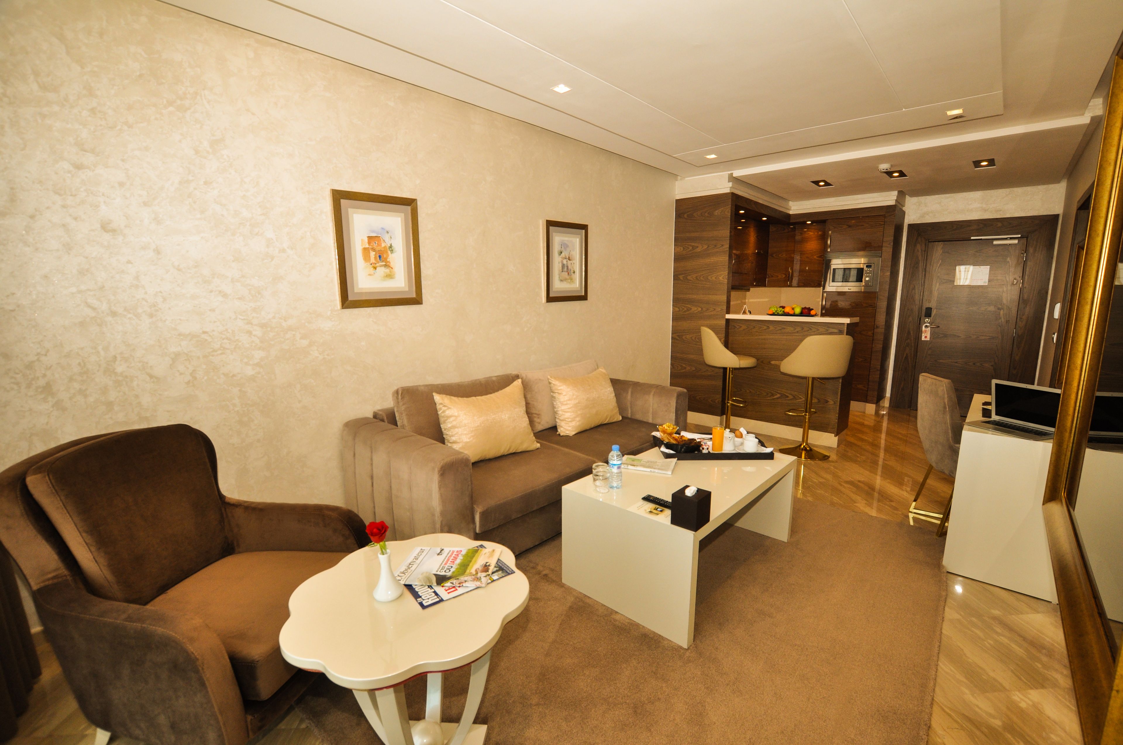 A modern living room with a brown sofa, a white coffee table, and a small table with a vase and magazines. There is a kitchen area with a microwave and a TV.