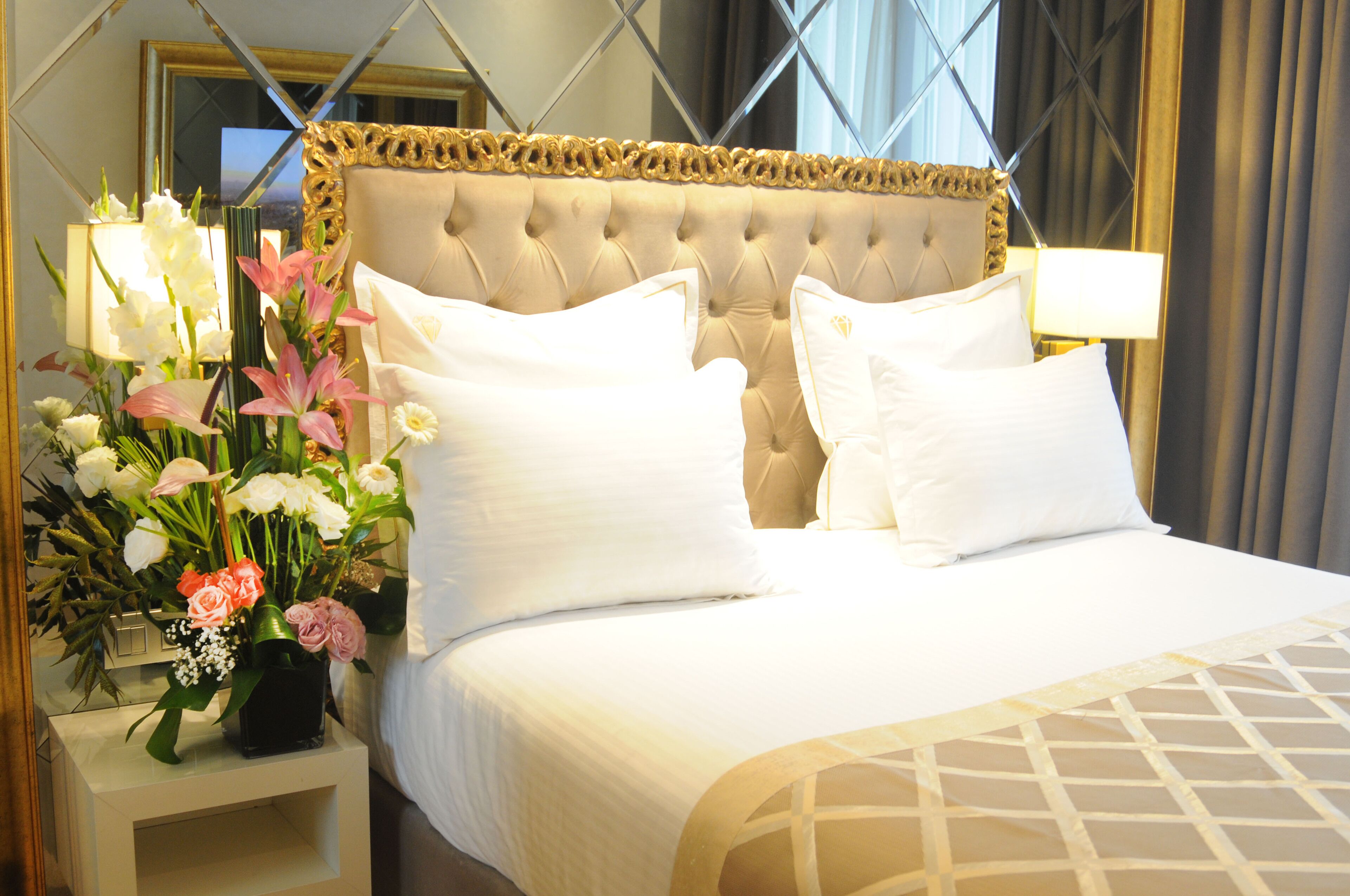 A hotel room with a tufted headboard, white bedding, and a floral arrangement on the nightstand.
