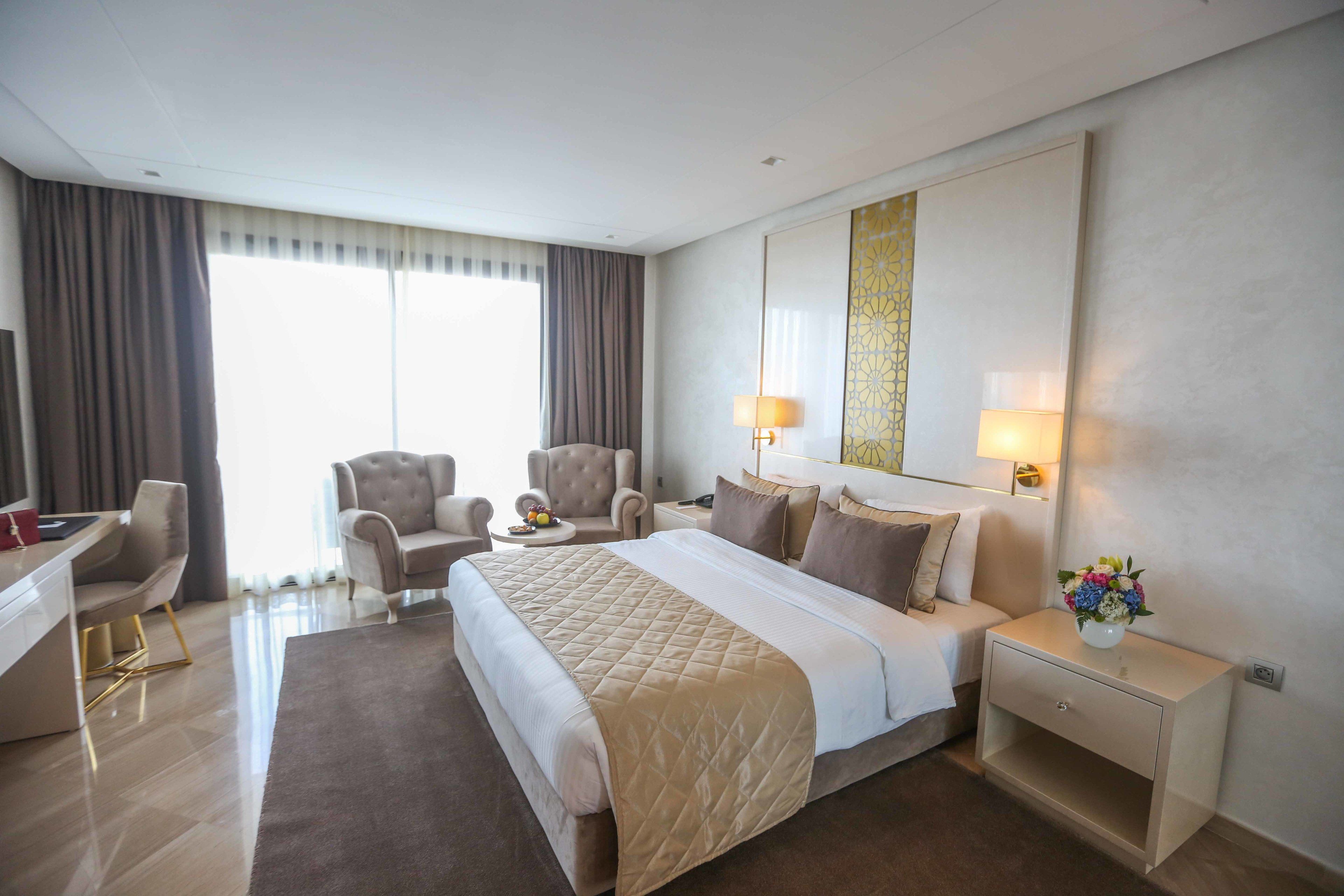 A hotel room with a large bed, two armchairs, a desk, and a nightstand with a lamp and vase of flowers.