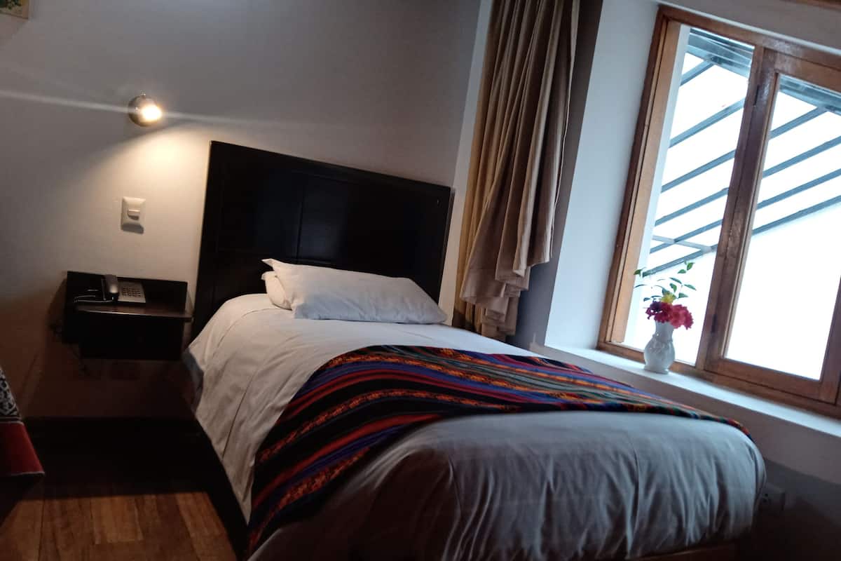 A bedroom with a bed, a bedside table with a phone, a window with curtains, and a plant on the windowsill.
