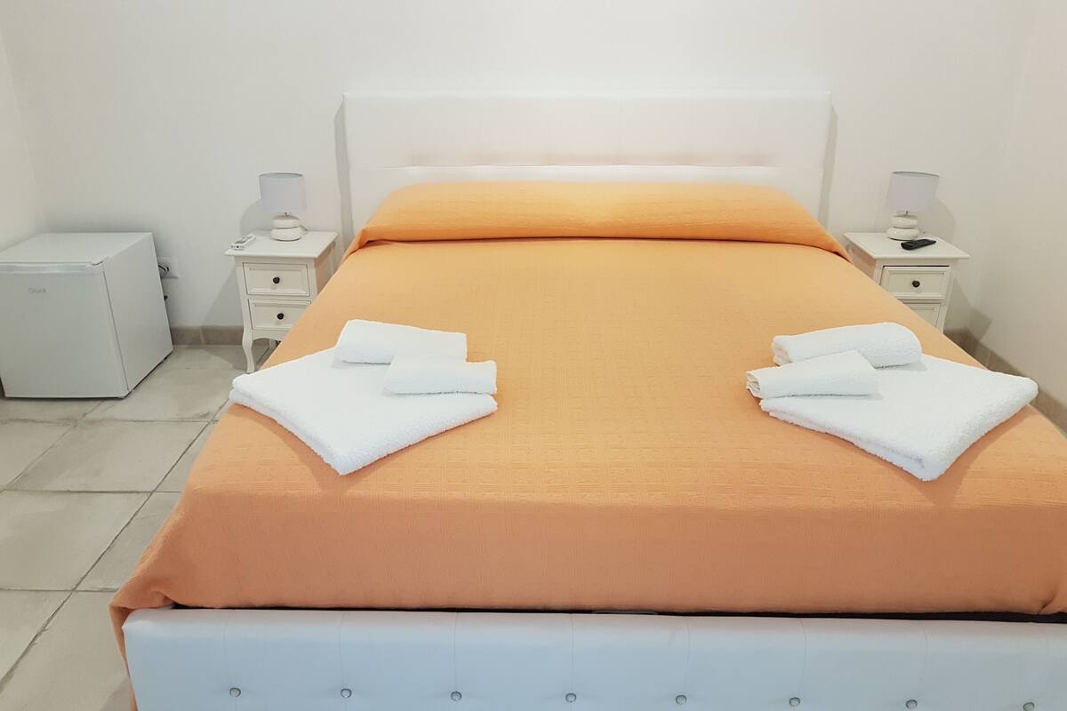 Ein ordentlich bezogenes Bett mit orangefarbener Bettdecke und weißen Kissen, zwei weiße Handtücher auf dem Bett, ein kleiner weißer Nachttisch mit Lampe sowie ein weiterer weißer Nachttisch mit Telefon und einem kleinen Gegenstand.