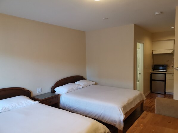 Habitación de hotel con dos camas, una mesa de madera, una kitchenette pequeña y una puerta que da a otra habitación.