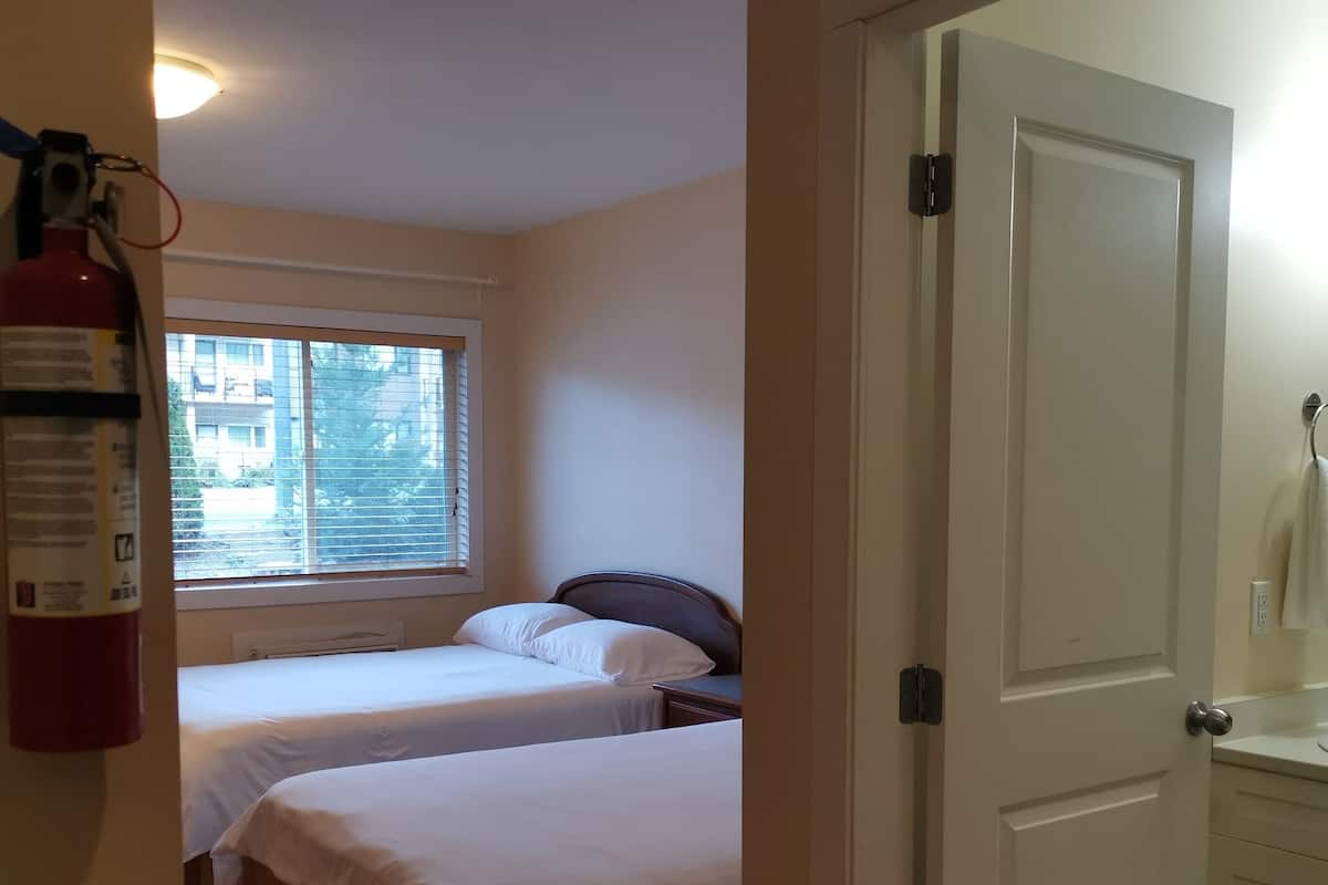 A bedroom with two beds, a window with blinds, a fire extinguisher, and a door leading to a bathroom.