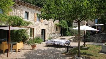 Un edificio de piedra con un patio de grava, asientos al aire libre y un árbol que da sombra.