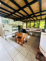 Una cocina con una mesa de madera y taburetes, suelo de baldosas y una ventana con vegetación al exterior.