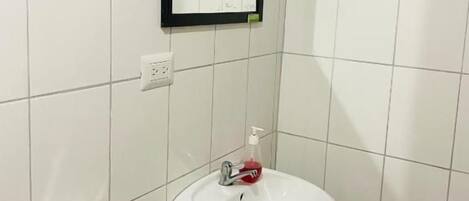 Un baño con lavamanos blanco, espejo, toallero y dispensador de papel higiénico.