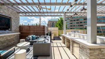 An outdoor patio with a pergola, seating, a TV, and a view of a building in the background.