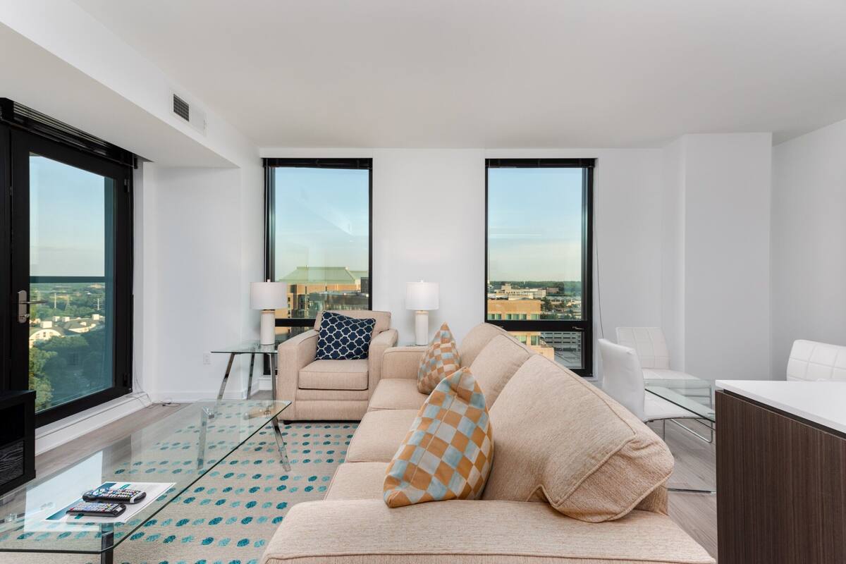 A modern living room with a beige sofa, a glass coffee table, and a view of the city through large windows.