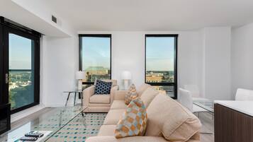 A modern living room with a beige sofa, a glass coffee table, and a view of the city through large windows.