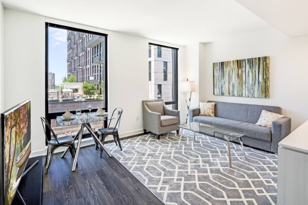 A modern living room with a dining area, large windows, a gray sofa, and a geometric patterned rug.