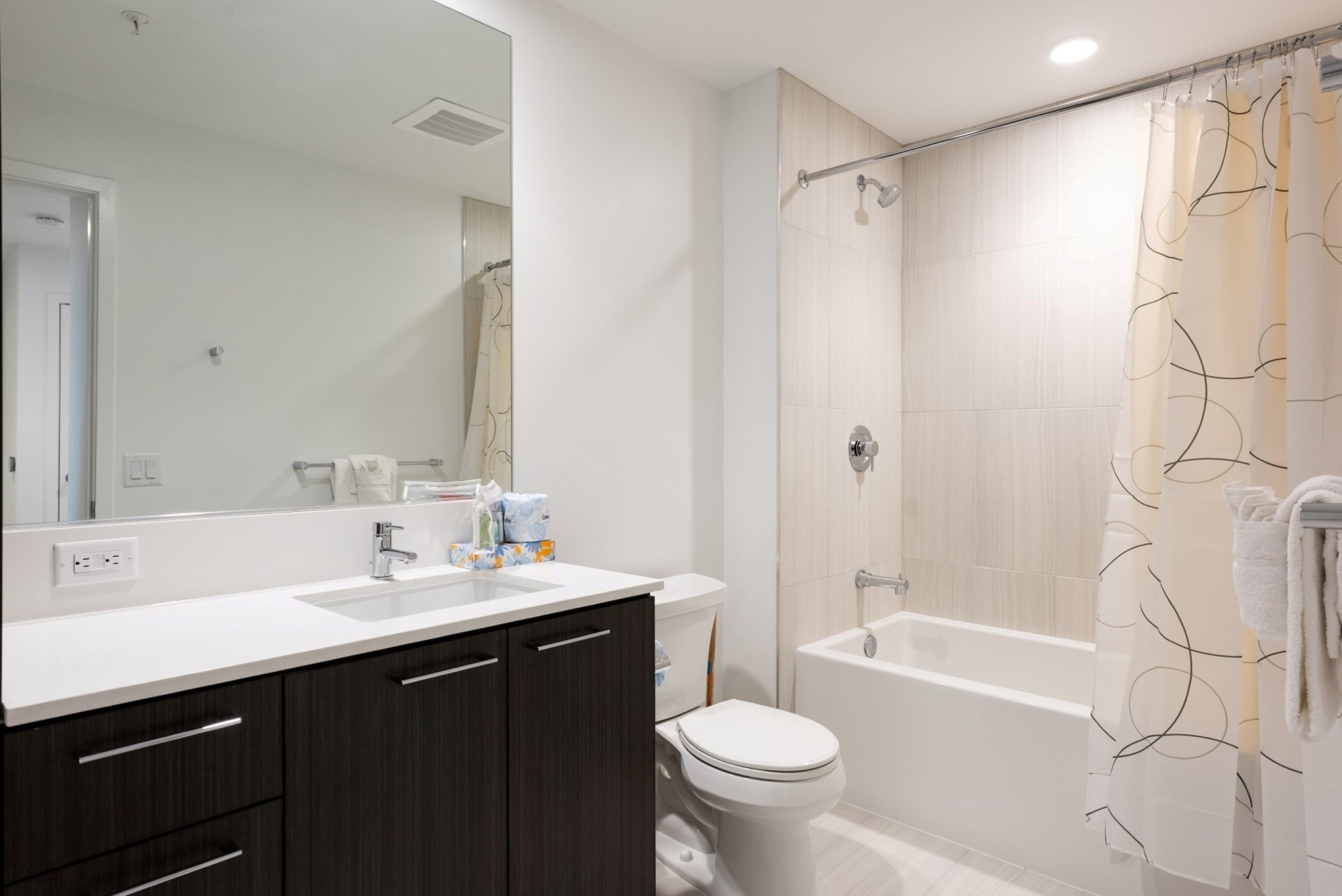 Un baño moderno con un lavabo blanco, muebles de madera oscura, un inodoro y una ducha a ras de suelo con cortina color beige.