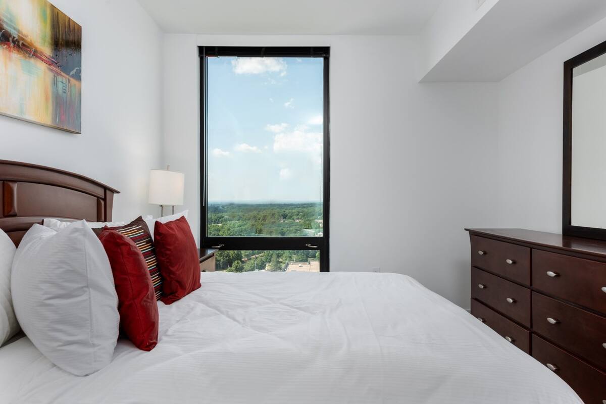 A bedroom with a large bed, a dresser, a window with a city view, and a painting on the wall.