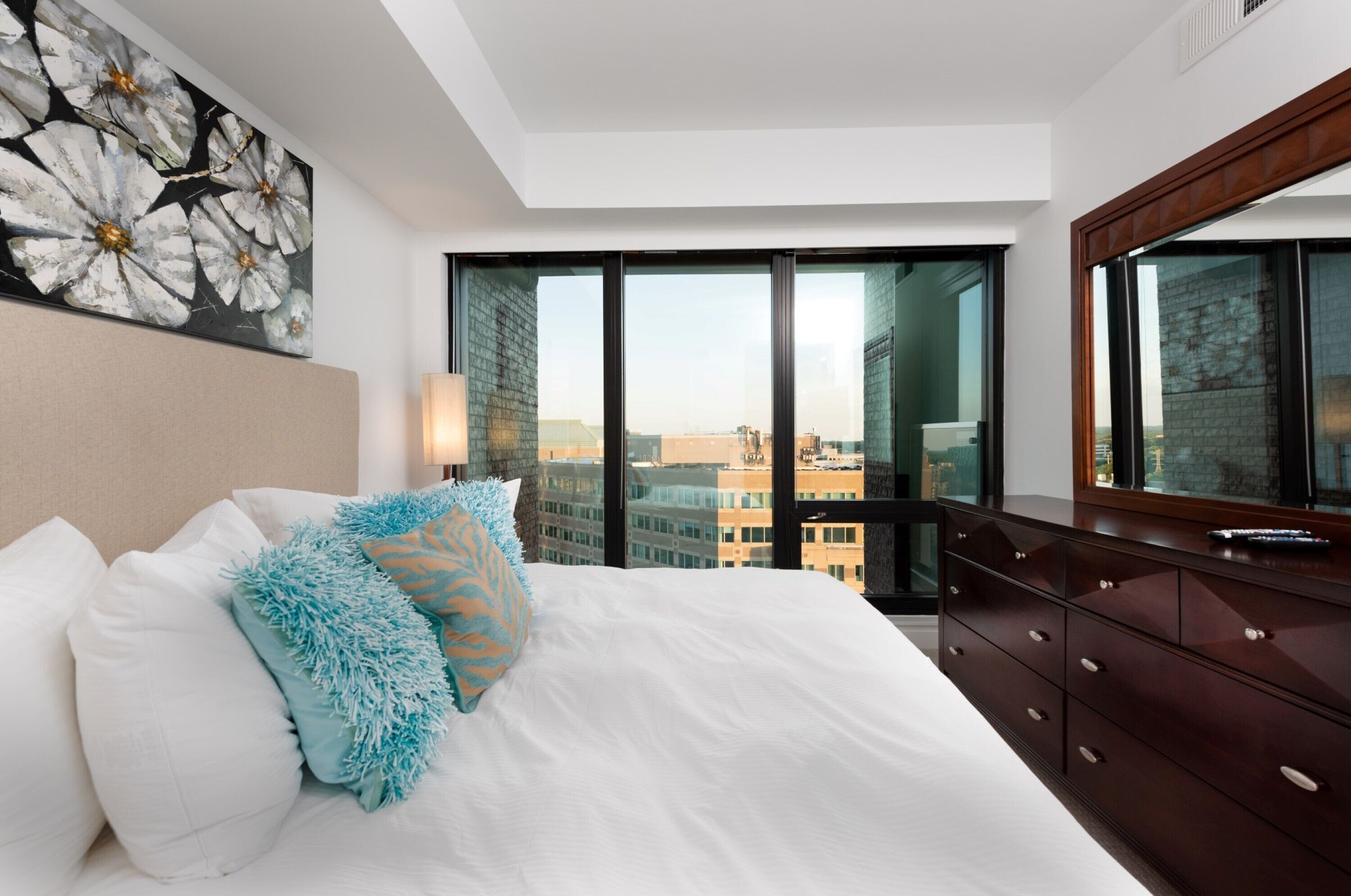 Une chambre d’hôtel avec un grand lit, une commode et une vue sur la ville grâce à de grandes fenêtres du sol au plafond.