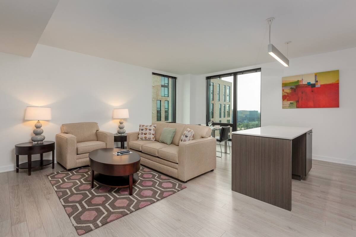 A modern living room with a sofa, coffee table, and a desk. There are two lamps on side tables, and a large window with a view of buildings.