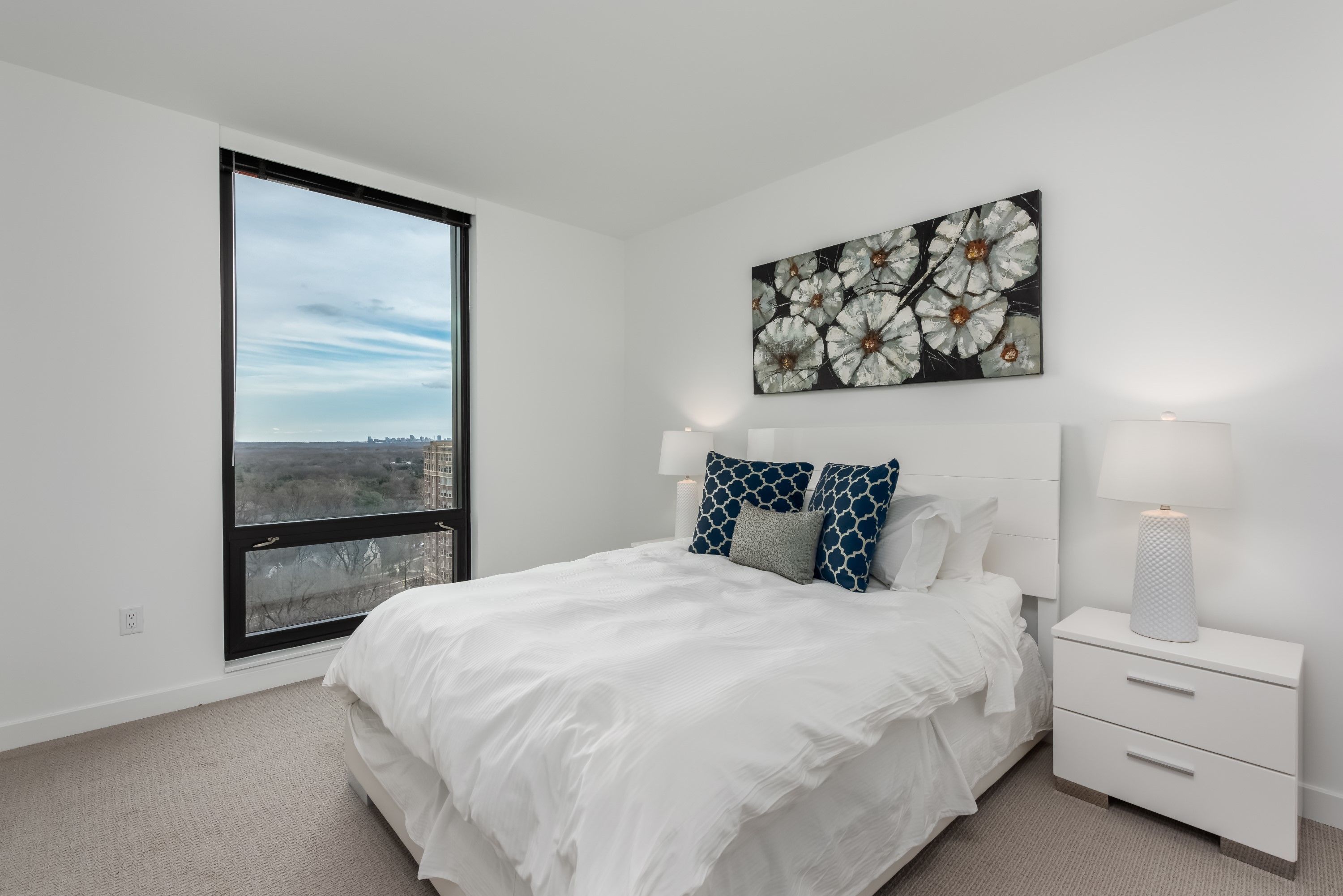 A bedroom with a large window, a bed with white bedding, a nightstand with a lamp, and a floral artwork on the wall.