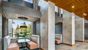 A modern interior with a seating area featuring cushioned benches and a glass table, surrounded by large concrete pillars and a wooden ceiling.