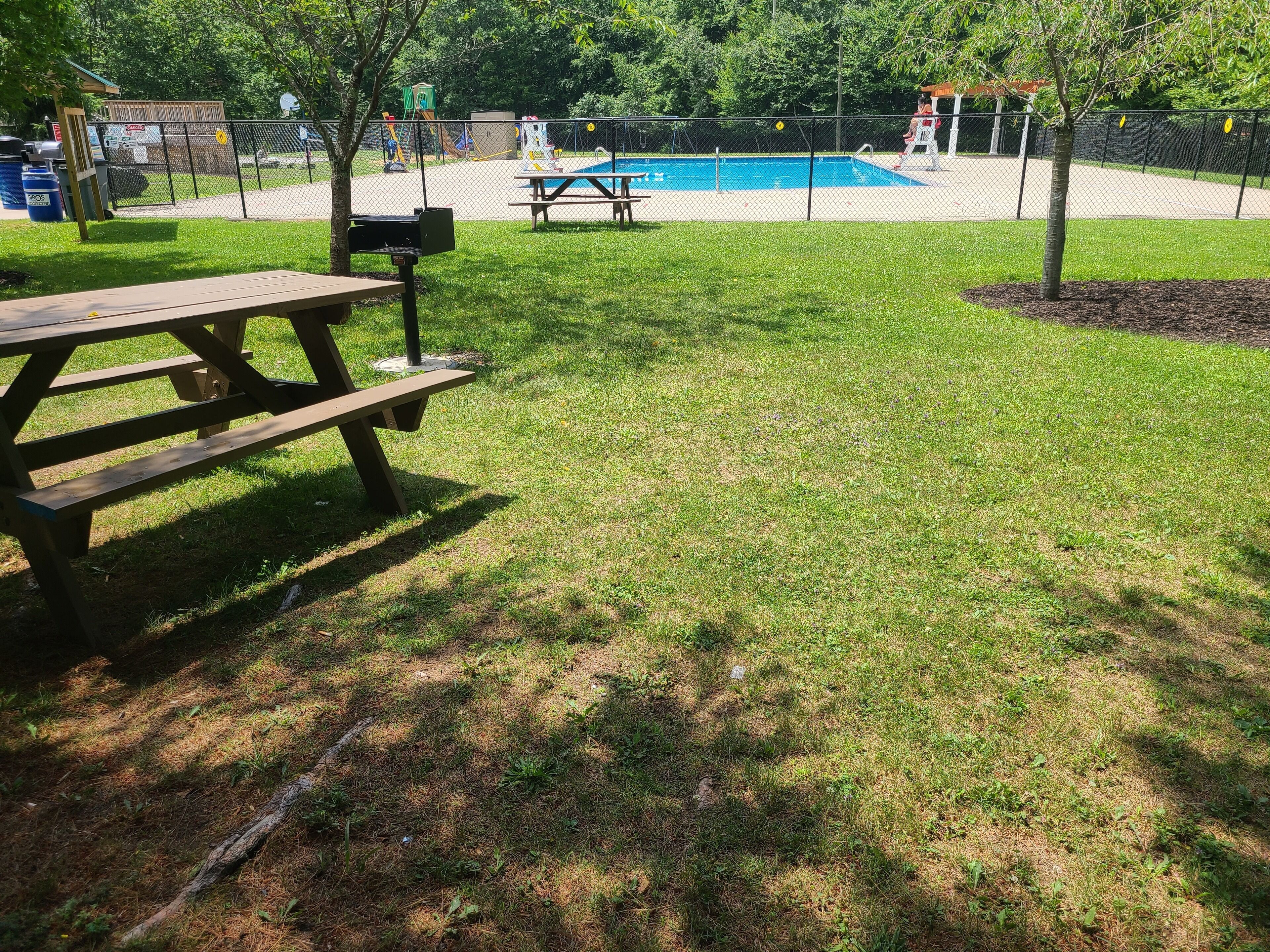 Un espace verdoyant avec une table de pique-nique, une piscine clôturée et, au fond, des jeux pour enfants.