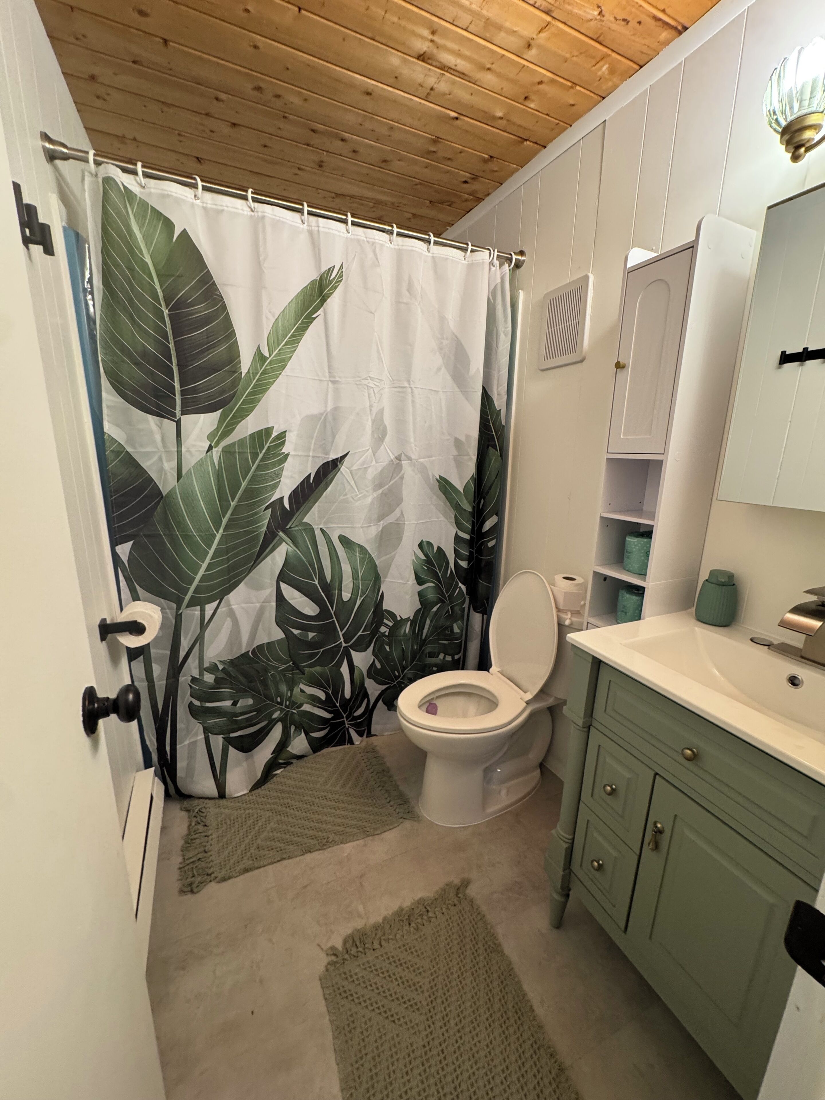 Une salle de bain comprenant des toilettes, un lavabo, un meuble de rangement, un rideau de douche orné d’un motif de feuilles et un plafond en bois.