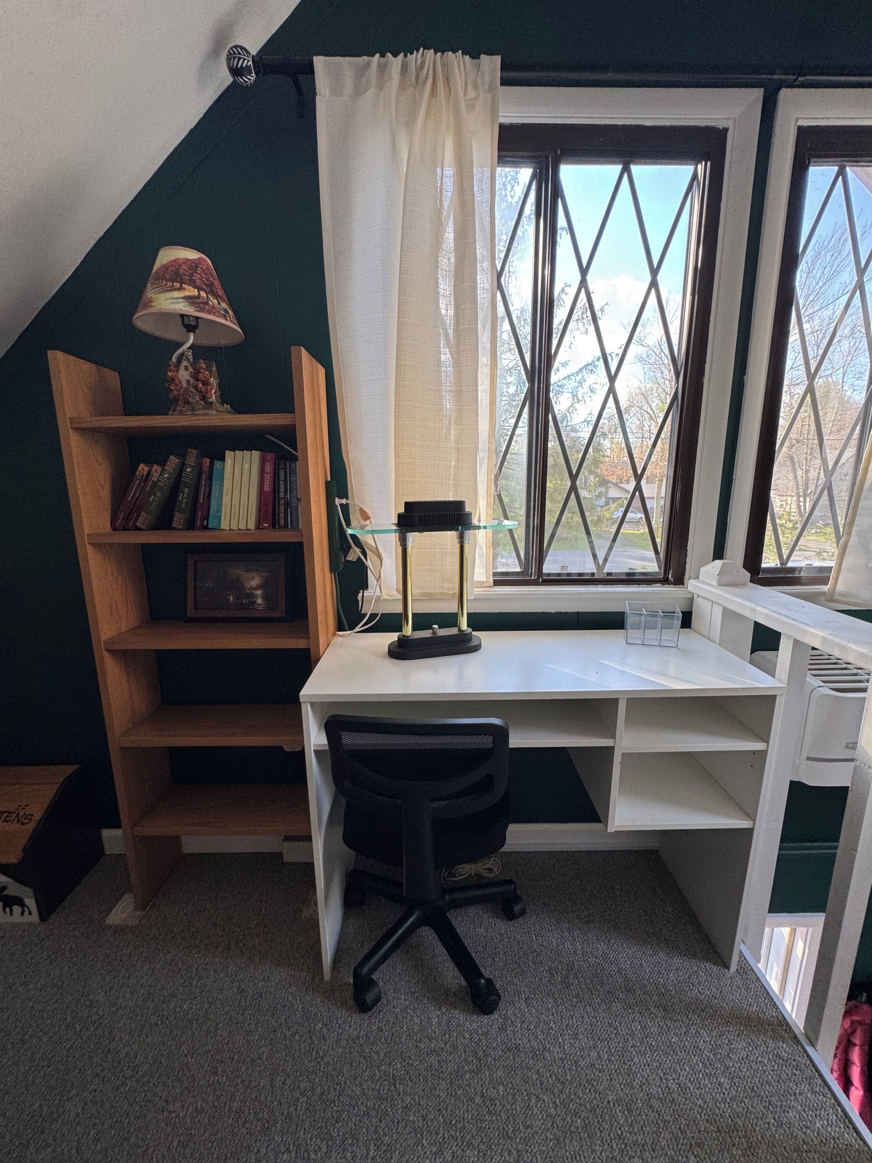 Una habitación con techo inclinado, un escritorio con silla, una estantería, una ventana con cortinas y una lámpara.