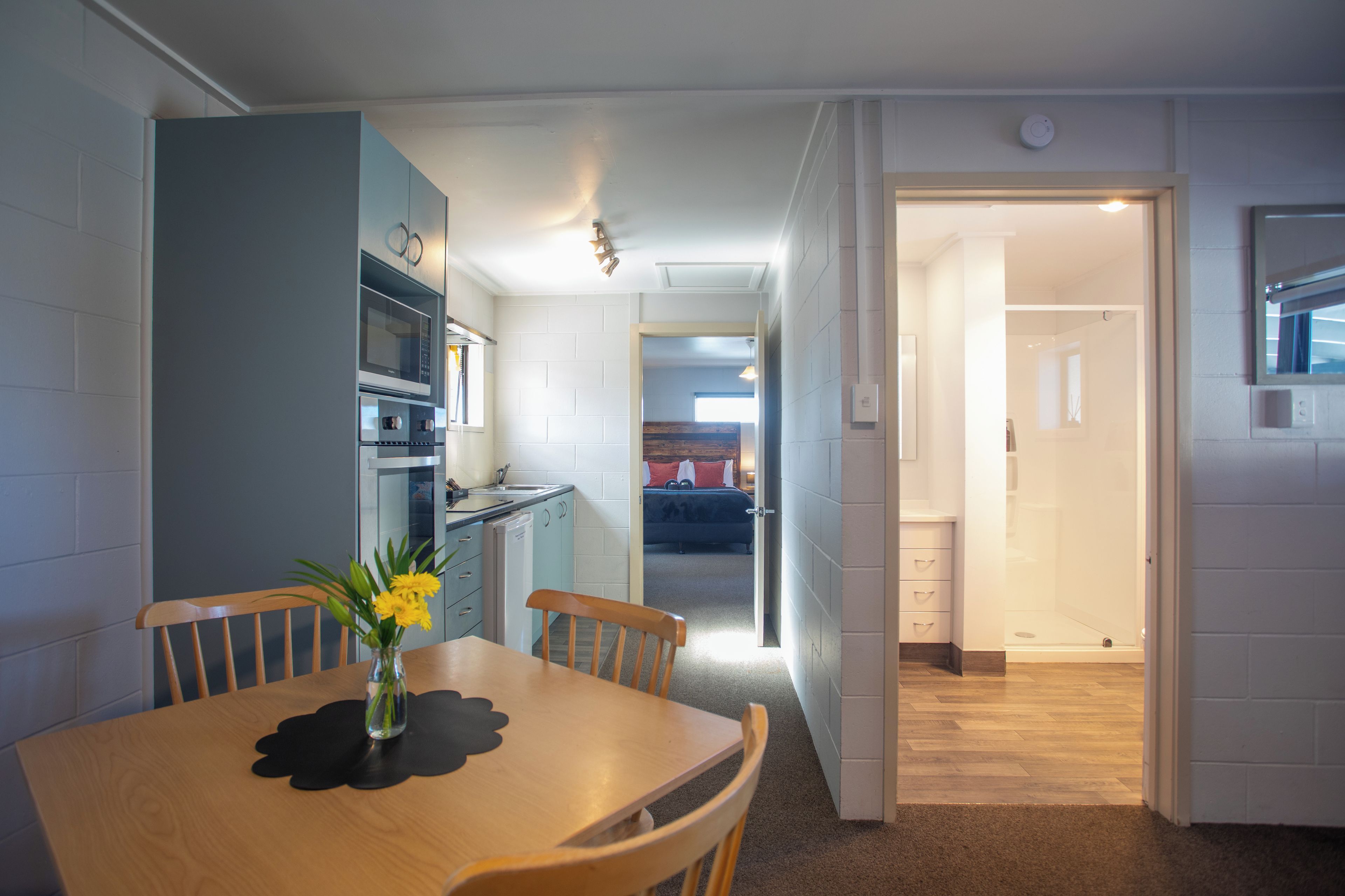 Cocina compacta con microondas, horno y lavamanos. Hay una mesa de comedor con sillas y un florero. Un pasillo conduce a un dormitorio con cama y ventana.