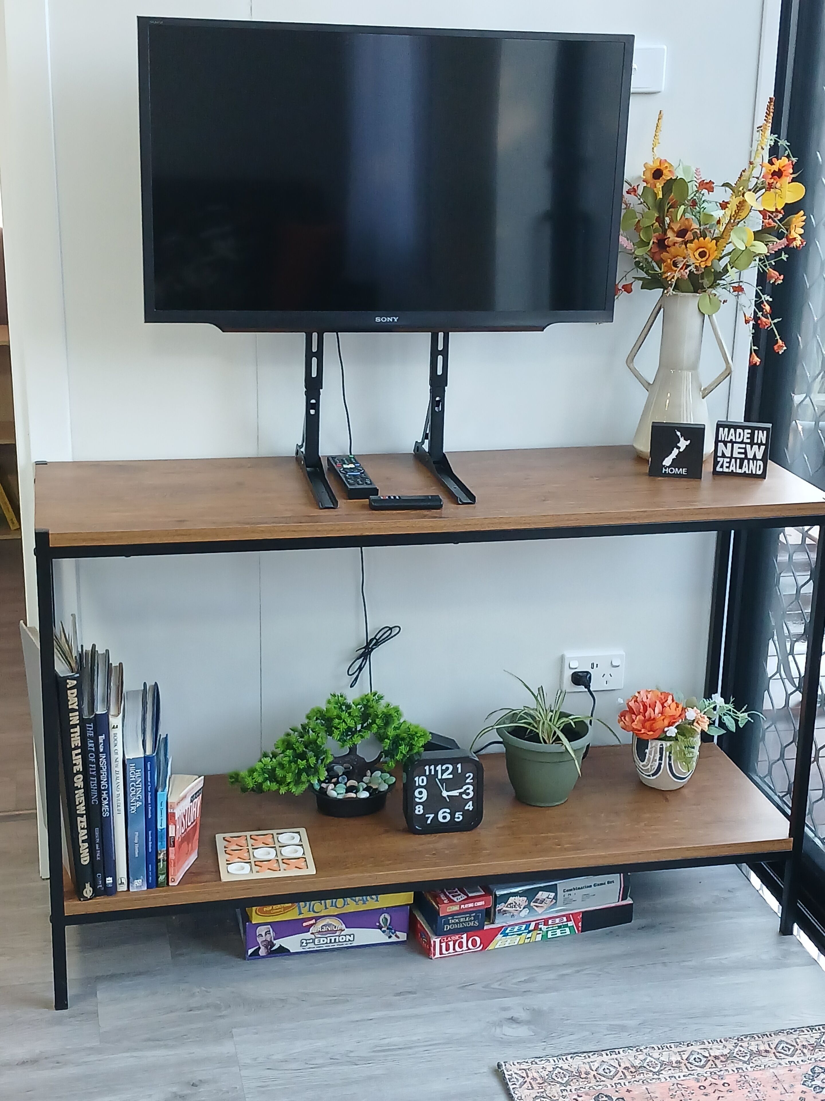 A flat-screen TV on a wooden stand with a remote control, a small sign indicating it's made in New Zealand, and a vase with flowers.