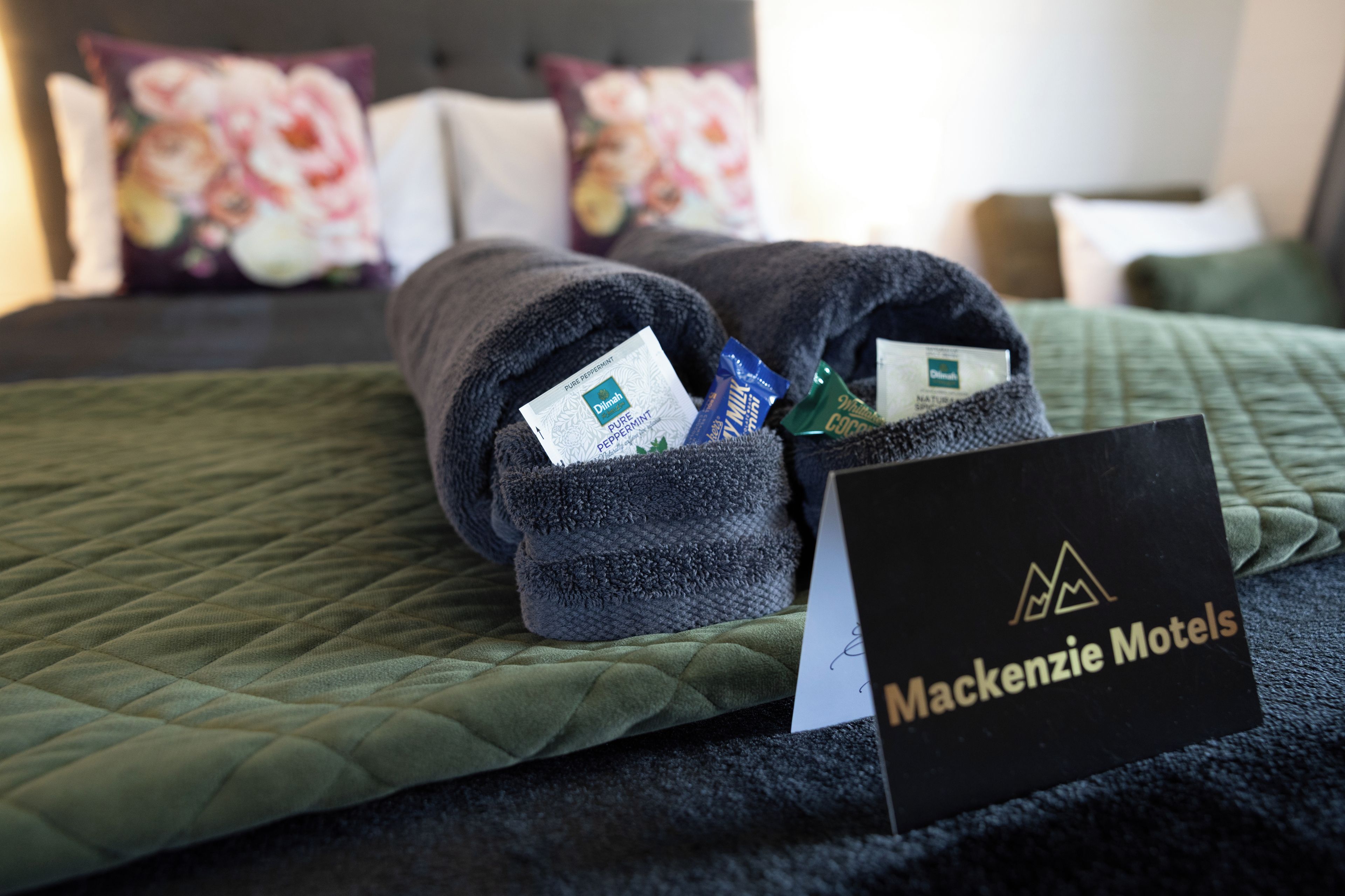 A neatly made bed with a green quilt, a sign for Mackenzie Motels, and a rolled towel with a packet of milk and a packet of pure peppermint.