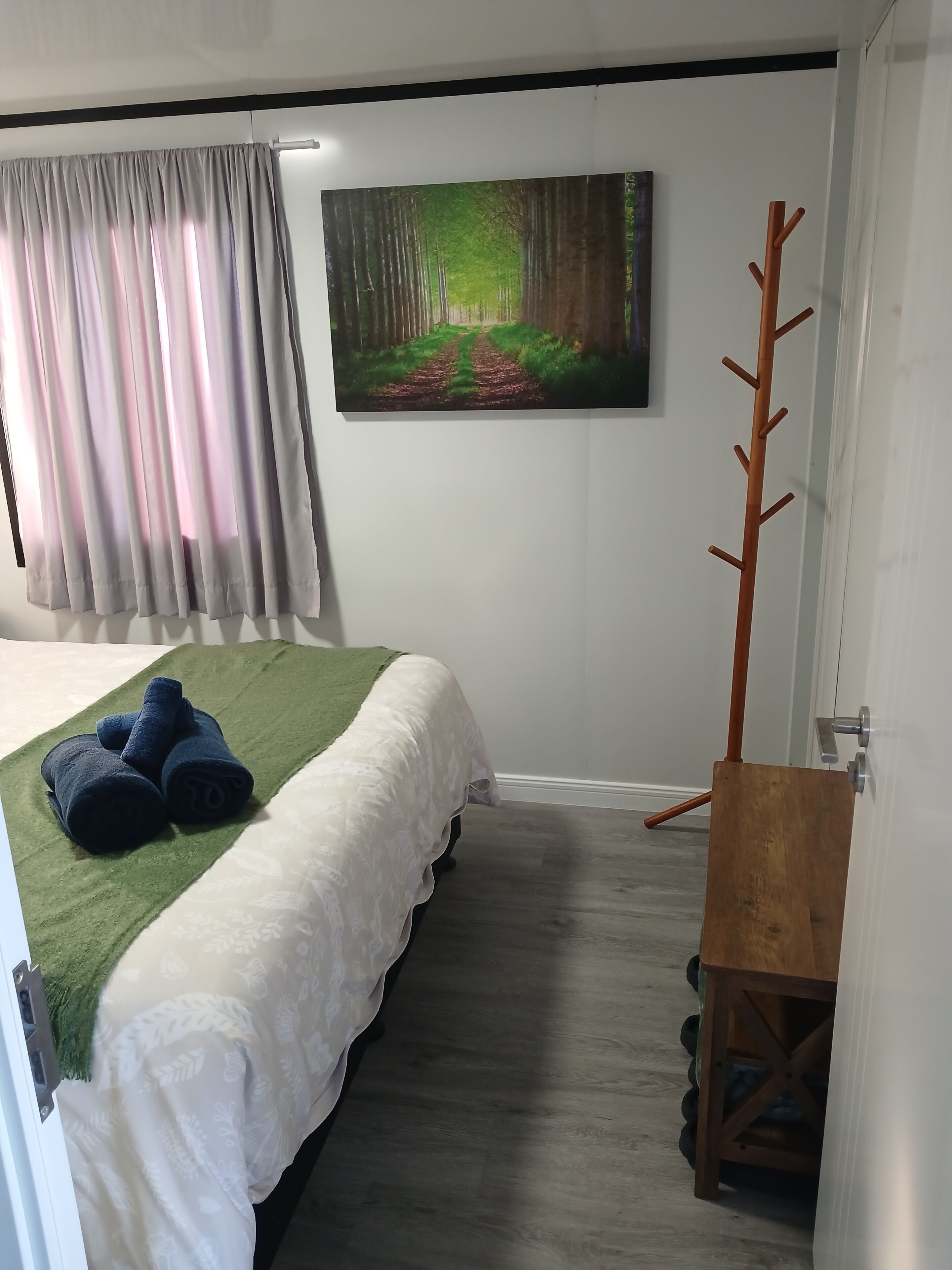 A bedroom with a bed, a painting of a forest, a coat rack, and a door.