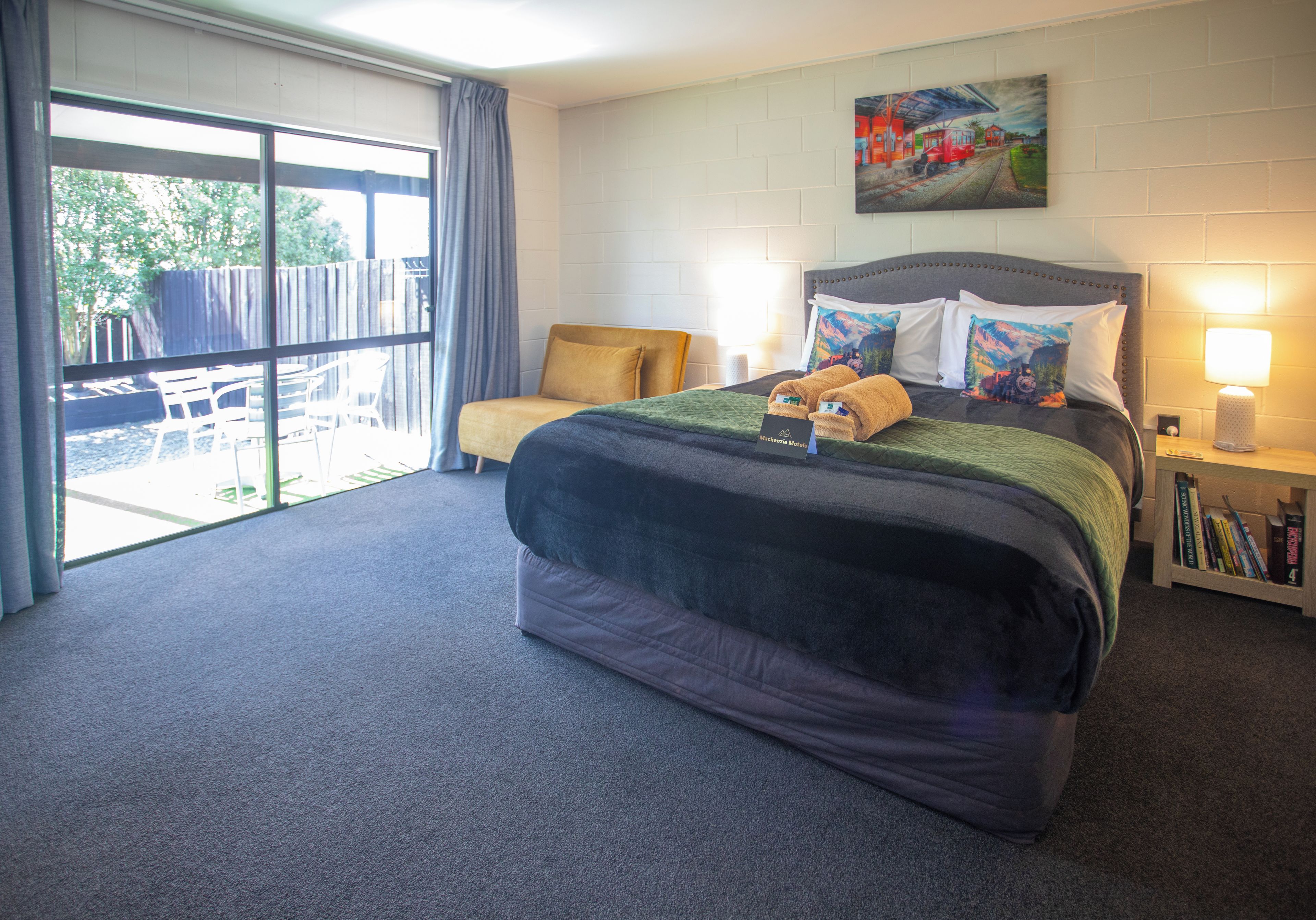A hotel room with a large bed, a side table with a lamp, a painting on the wall, and a view of the outdoors through a sliding door.