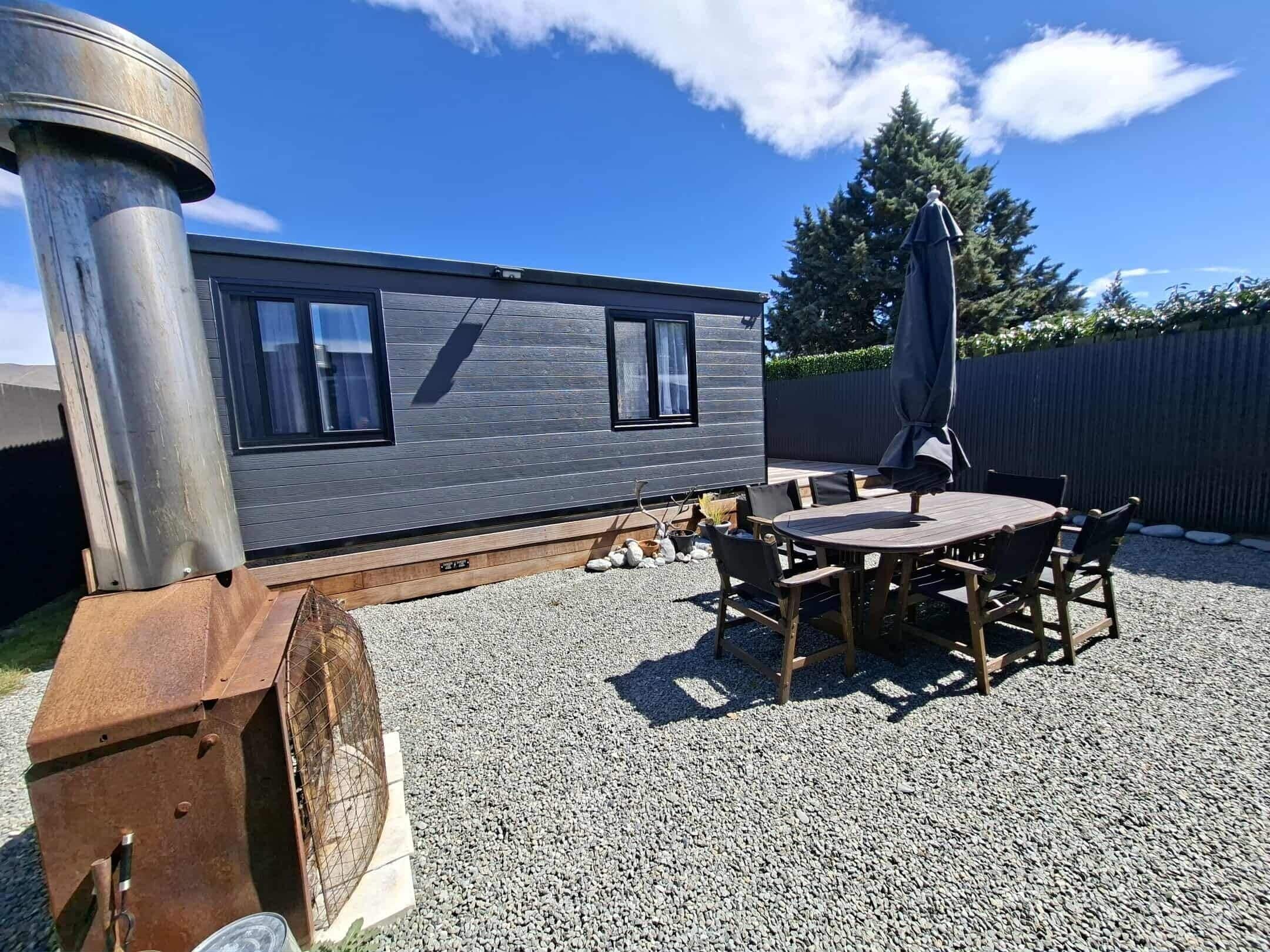 A modern, compact mobile home with a gravel patio, a wooden table and chairs, and a large umbrella.