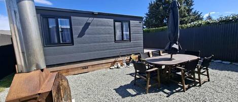 A modern, compact mobile home with a gravel patio, a wooden table and chairs, and a large umbrella.