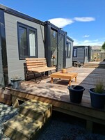 Una casa móvil moderna con un patio que incluye un banco de madera, una mesa y plantas en macetas.