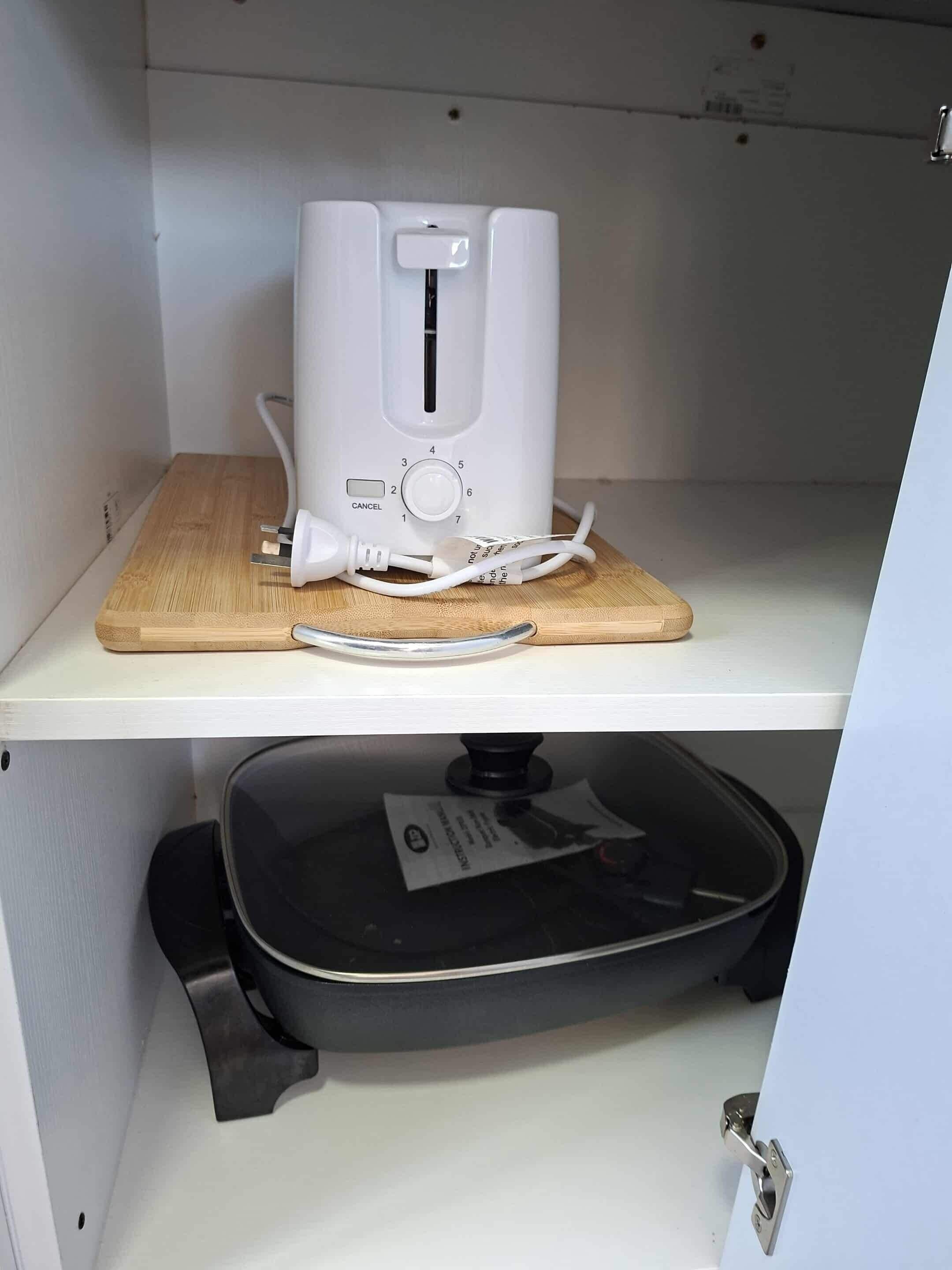Un tostador blanco sobre una tabla de madera en un estante blanco, con un electrodoméstico negro debajo.