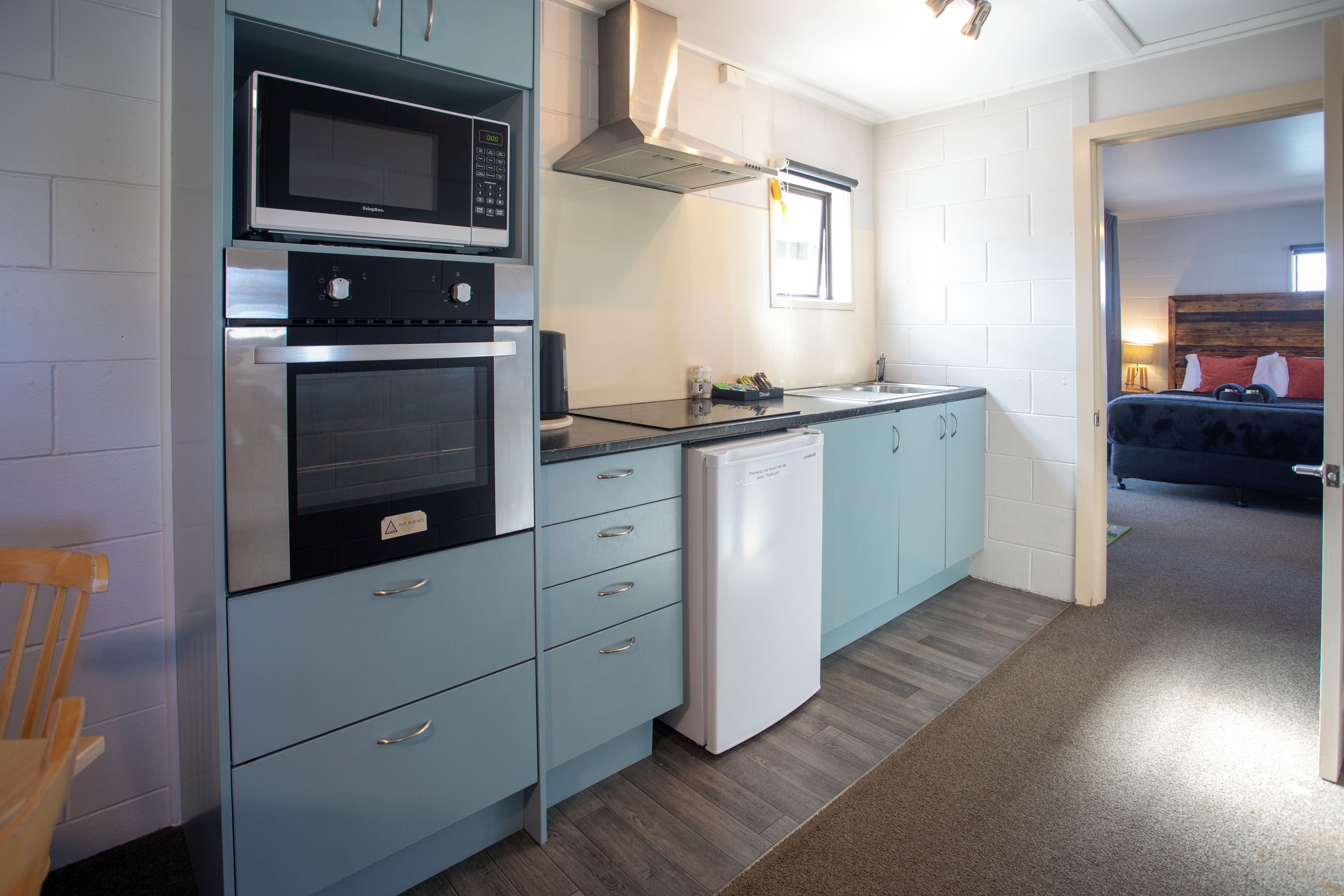 Cocina compacta equipada con microondas, horno y lavavajillas, armarios azul claro y una silla de madera.