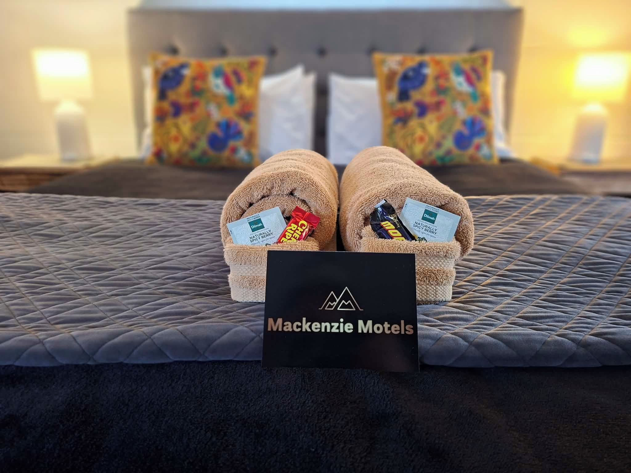A bed with a dark quilted bedspread, two pillows with colorful patterns, and a sign indicating the hotel name 'Mackenzie Hotels'.