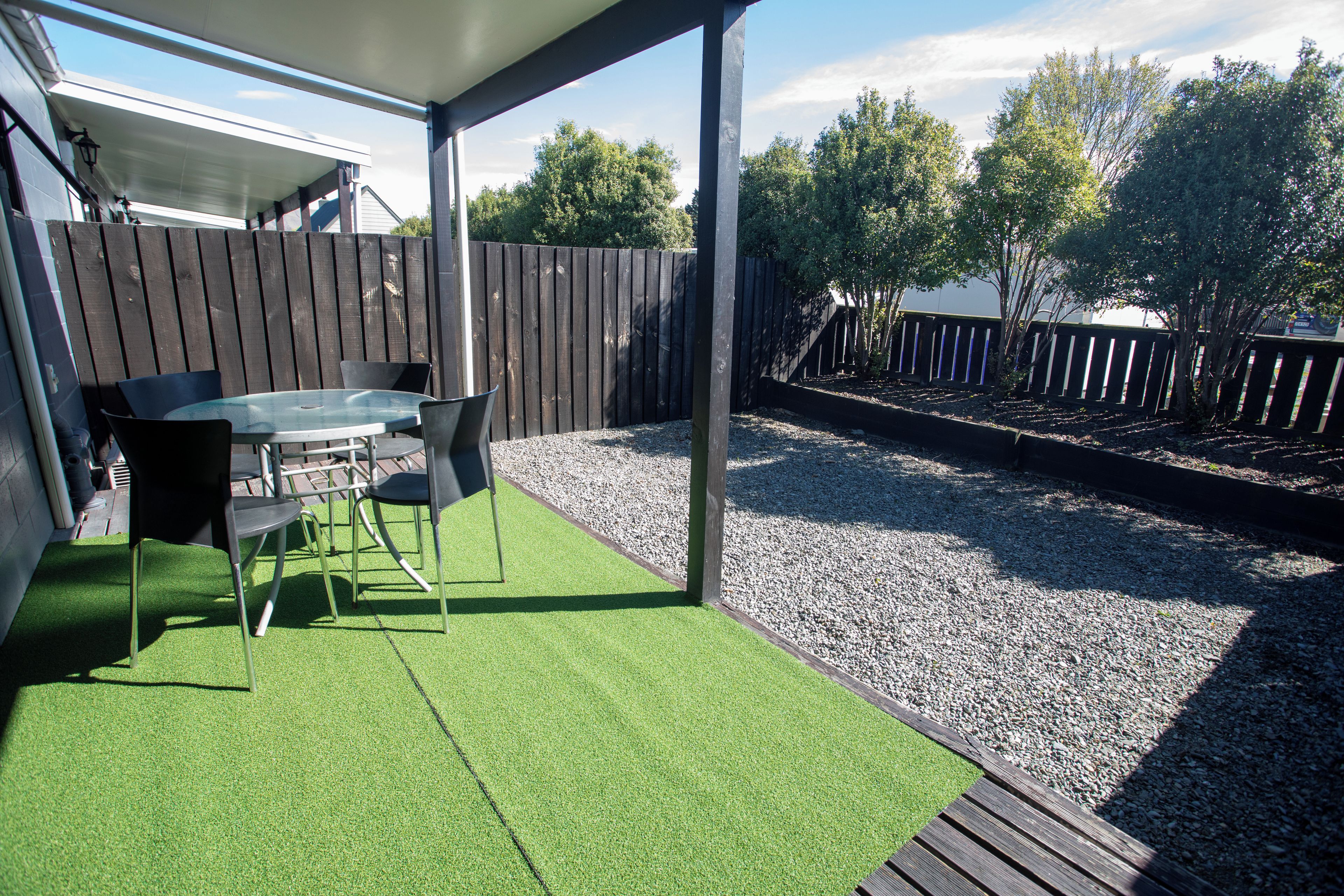 Un patio al aire libre con una mesa y sillas, rodeado por una cerca de madera y una zona de grava.