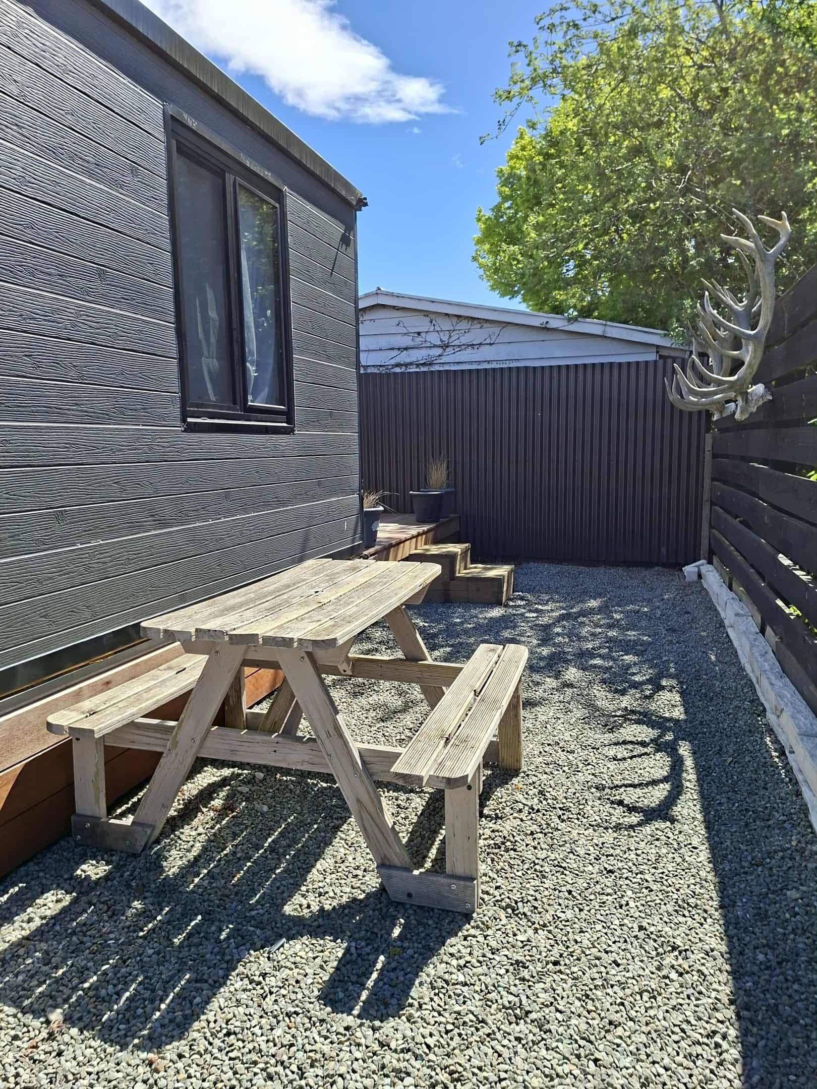 A wooden deck with picnic tables, a corrugated metal fence, and a building with a window.