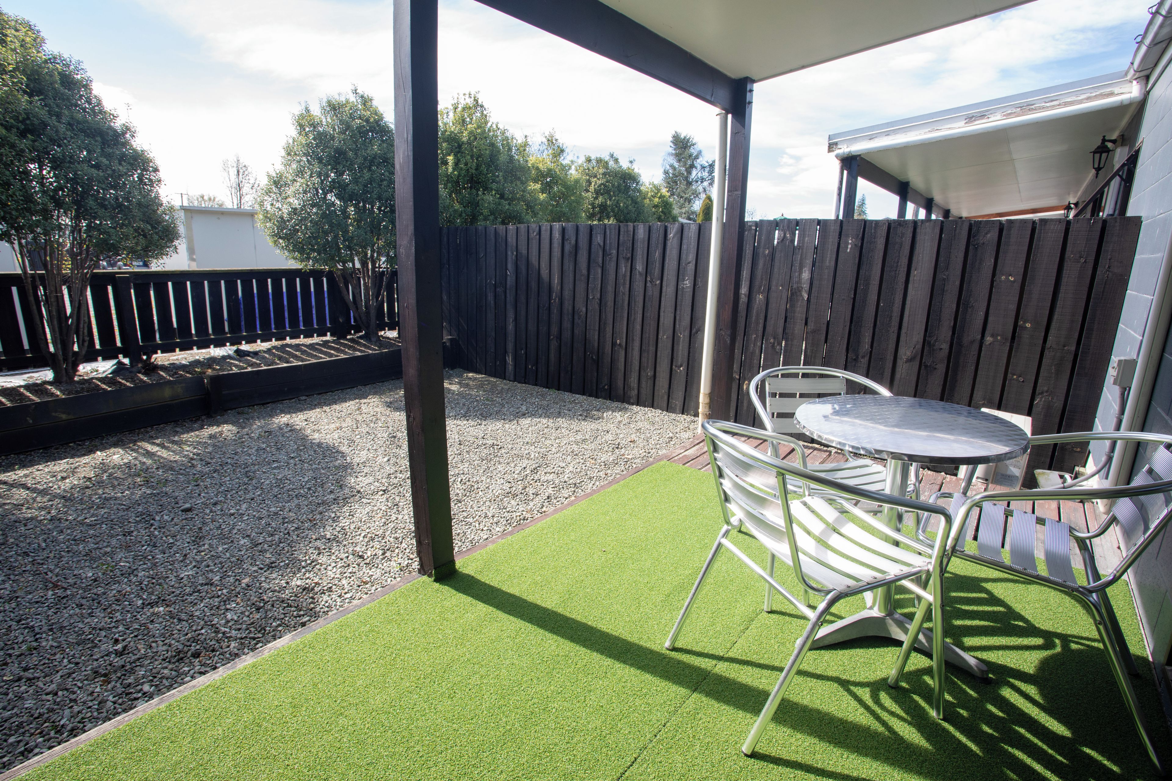 Een terras met kunstgras, een metalen tafel en stoelen, een houten schutting en een grindpad.