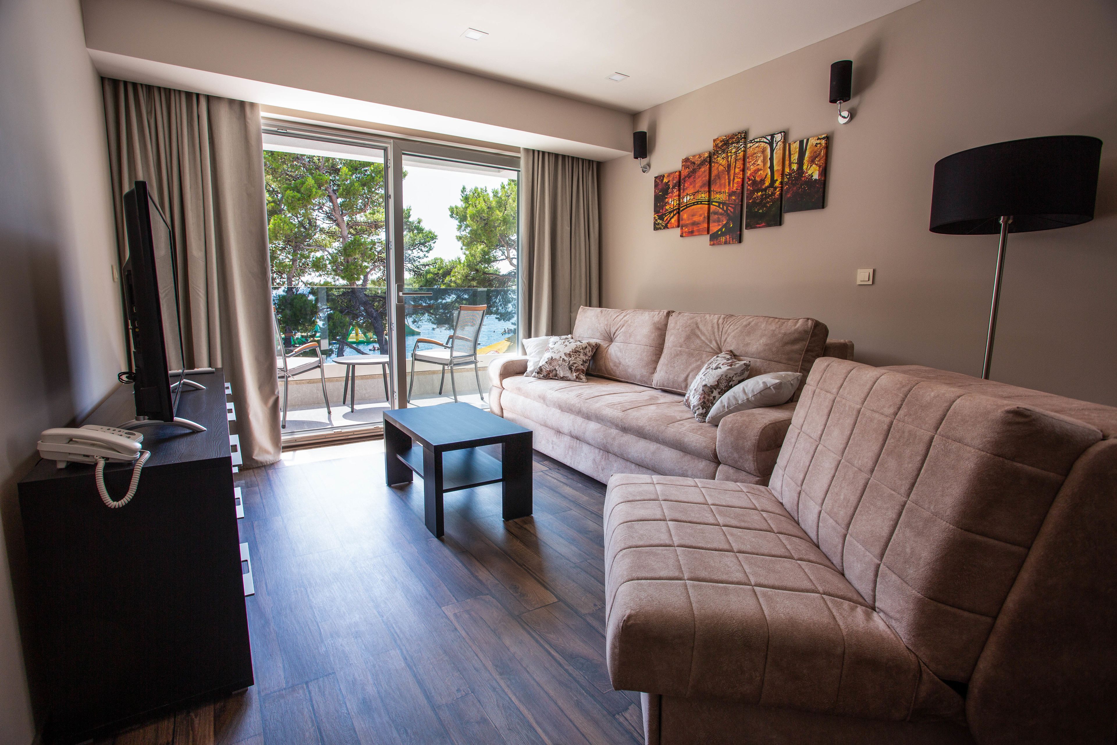 Ein modernes Wohnzimmer mit einer Couch, einem Couchtisch und einem Fernseher auf einem Schrank. Ein Balkon mit Stuhl und Tisch ist vorhanden.
