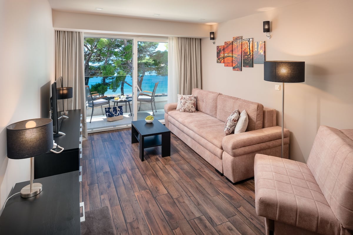 A modern living room with a sofa, a coffee table, and a TV. There is a balcony with a view of the sea.