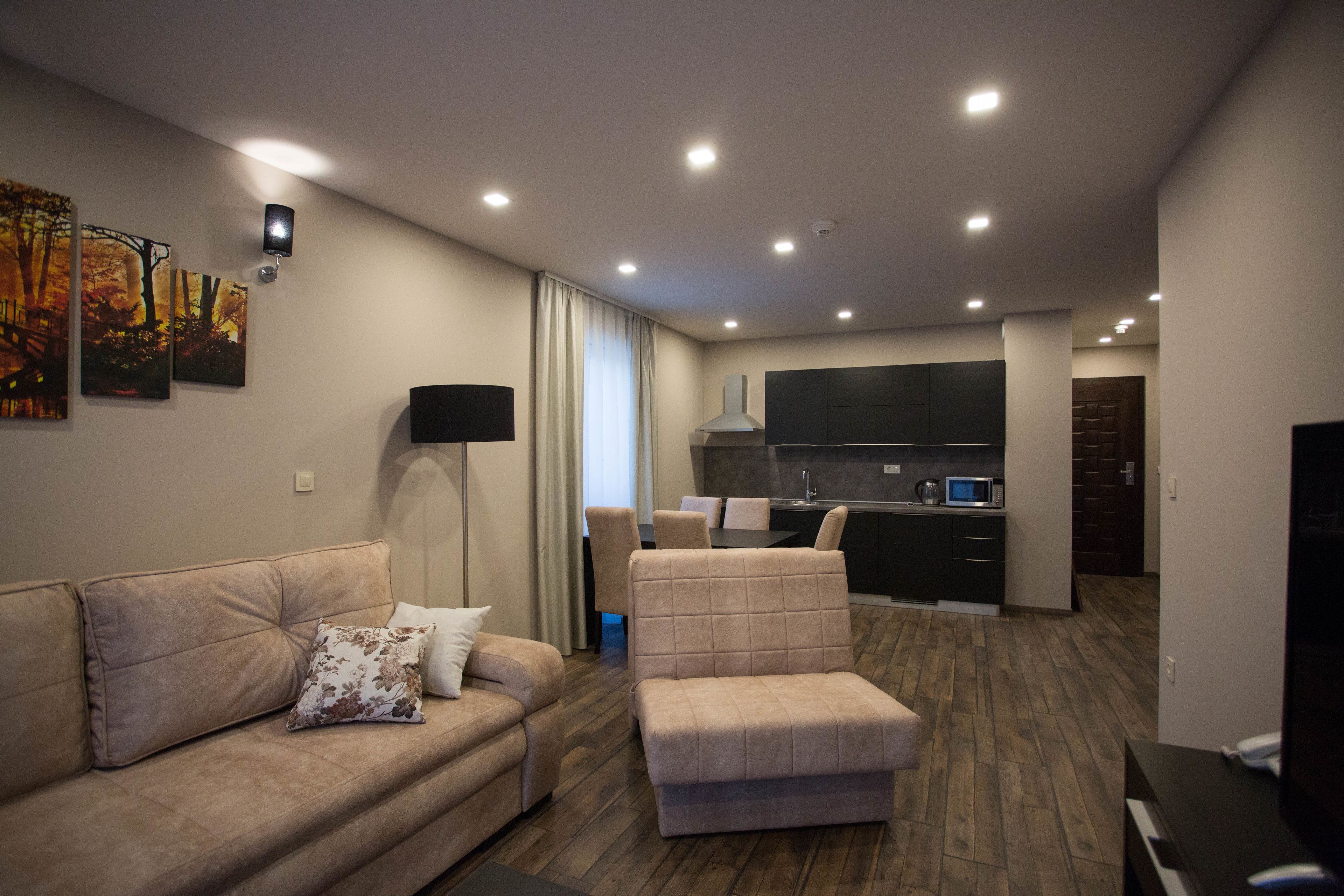 Una sala de estar moderna con un sofá, un reposapiés y un televisor de pantalla plana. Se ve un área de cocina con un microondas y armarios al fondo.