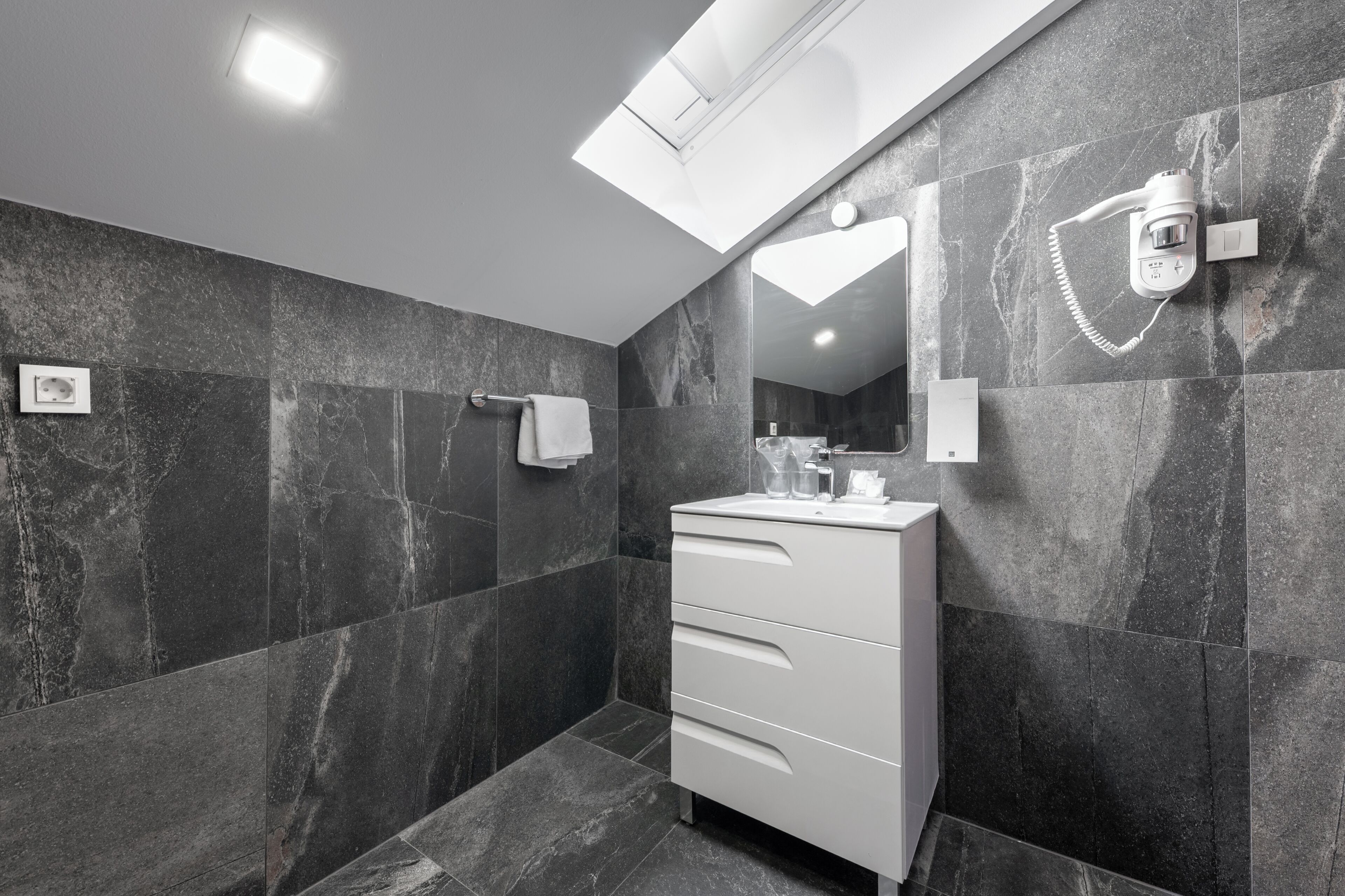 A modern bathroom with dark marble walls, a skylight, a white vanity, and a towel rack.