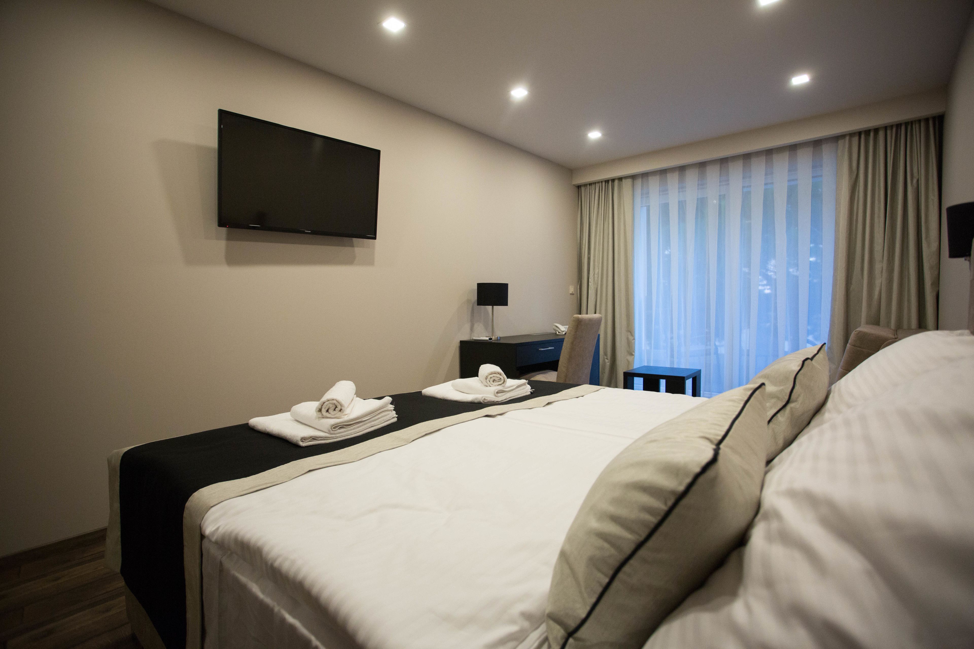 A hotel room with a bed, a flat-screen TV mounted on the wall, a desk with a lamp, and a window with curtains.