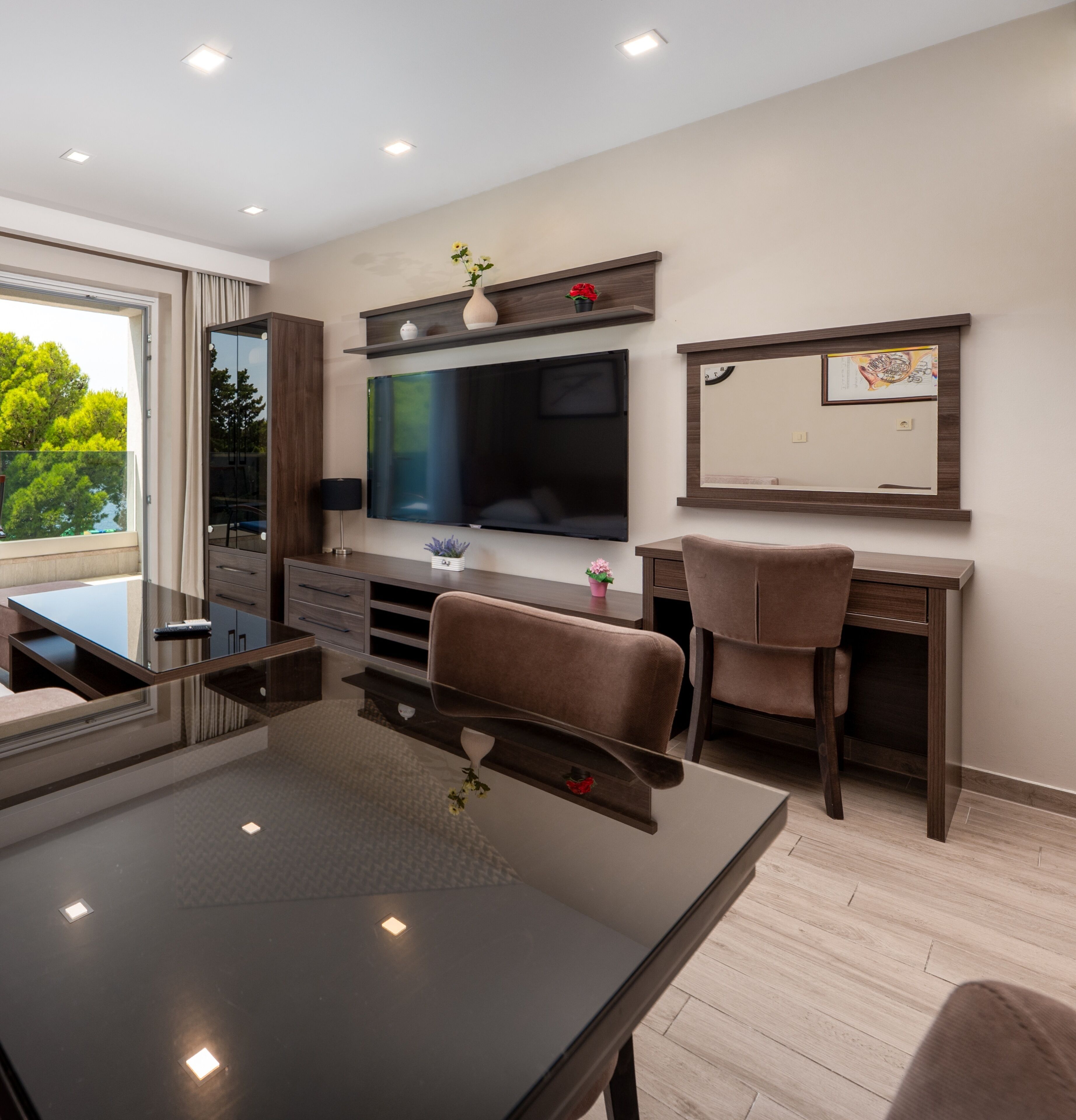 A modern hotel room with a large flat-screen TV, a dining table, two chairs, a desk, and a large window offering a view of trees.