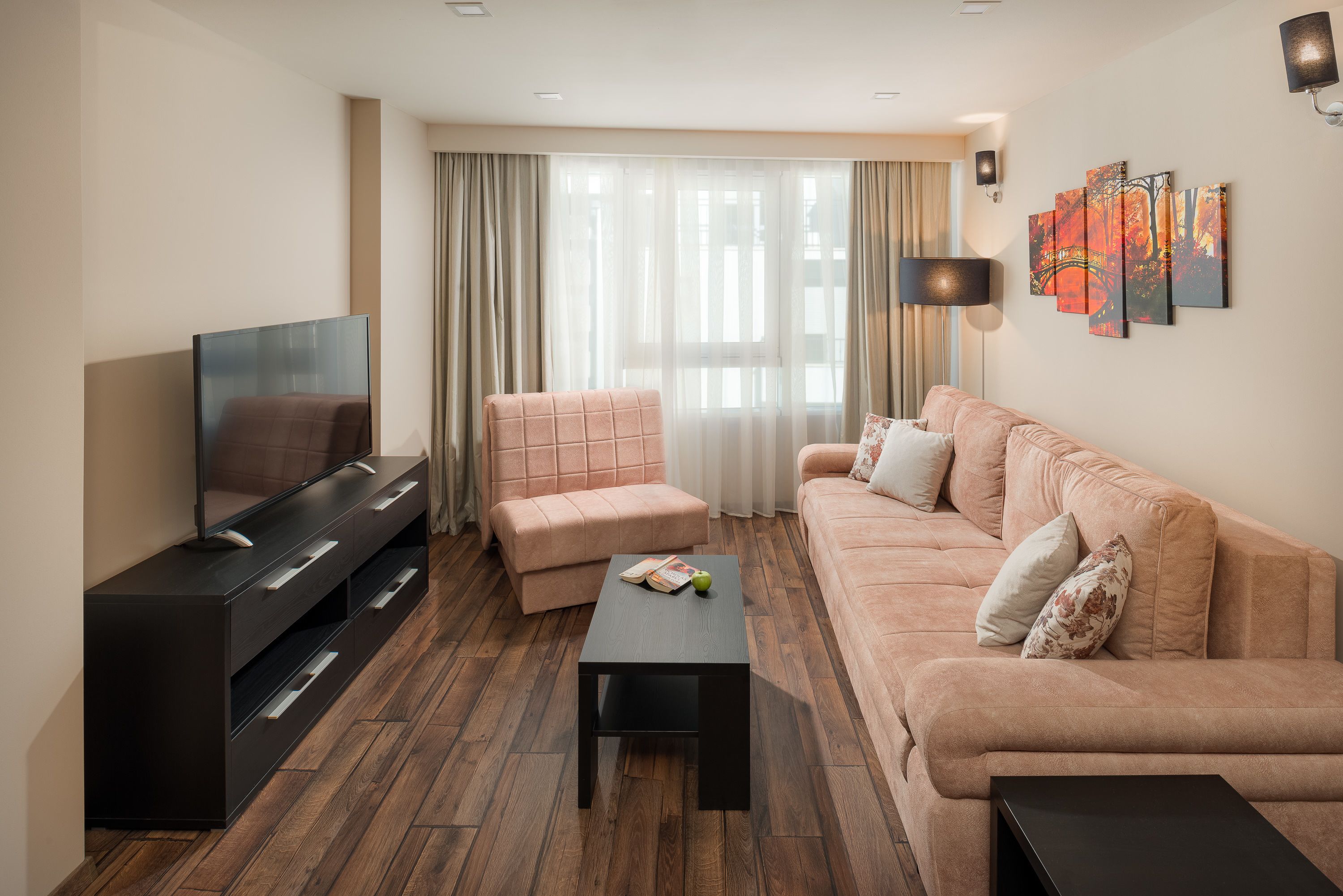 A modern living room with a flat-screen TV, a sofa, a loveseat, a coffee table, and a large window with sheer curtains.