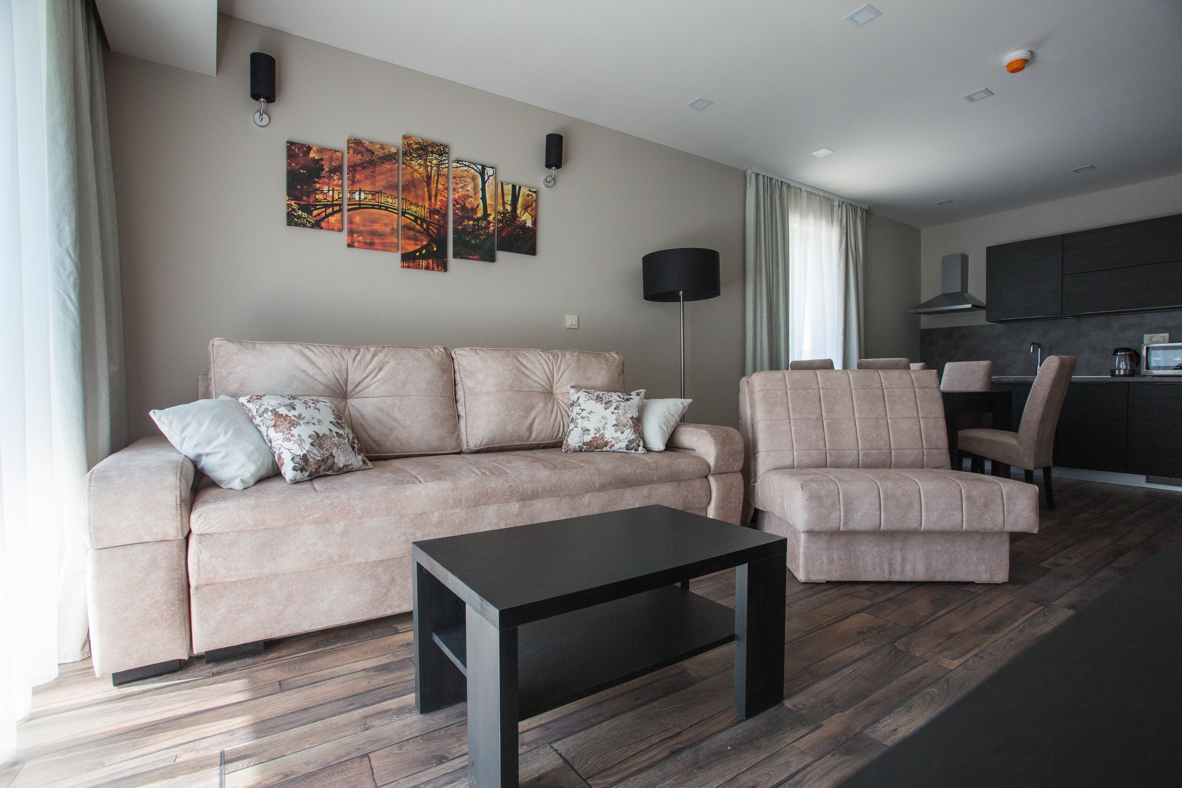 A modern living room with a sofa, armchair, and coffee table. There are wall art pieces and a kitchen area visible in the background.