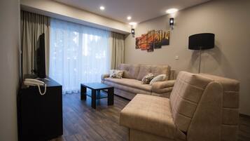 Una sala de estar moderna con un sofá, un reposapiés y una mesita. Hay un televisor de pantalla plana sobre un mueble, una lámpara y una obra de arte en la pared.