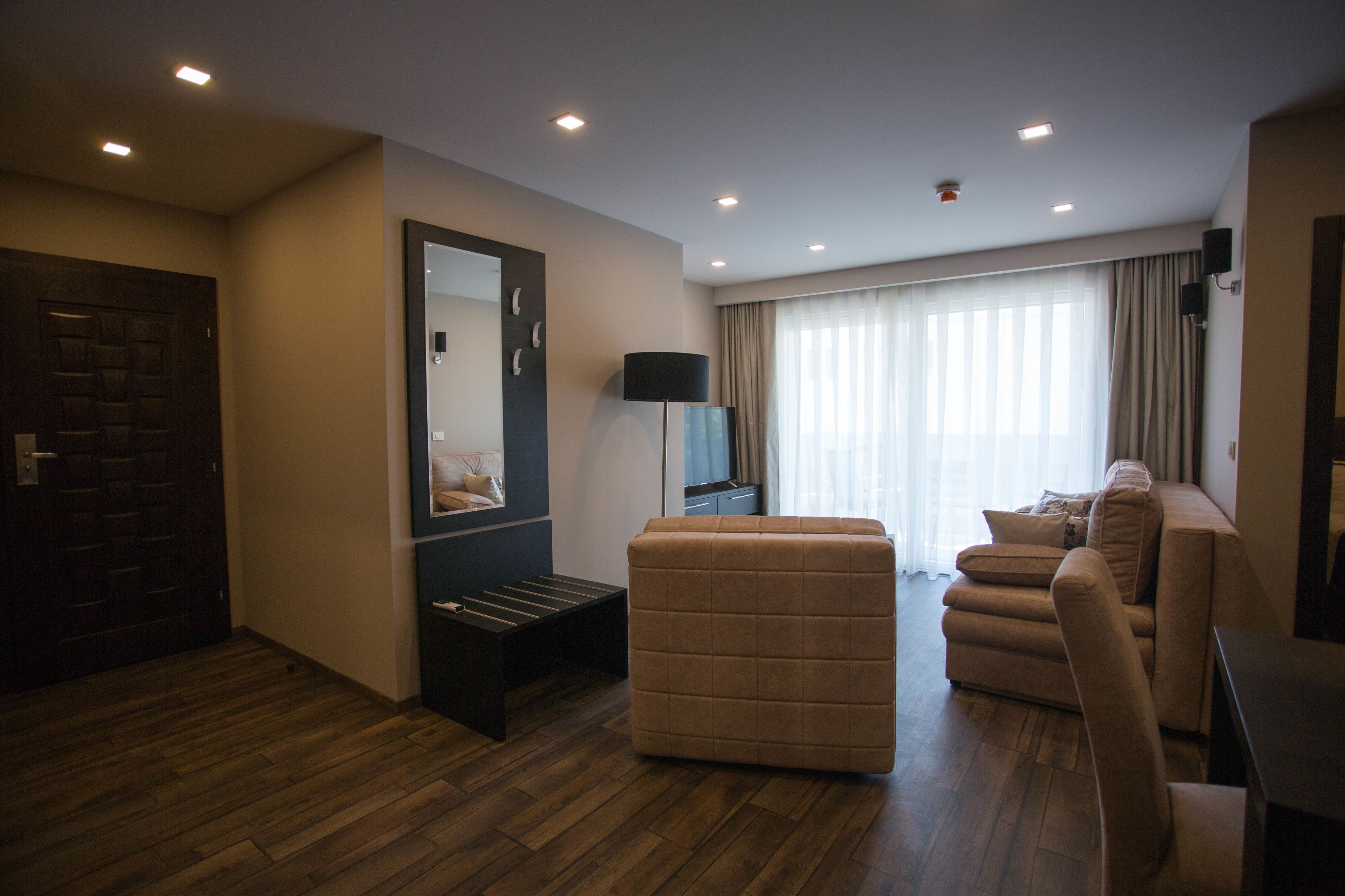 Una habitación de hotel moderna con piso de madera, un sofá, una silla, una mesita, un espejo, una lámpara y un televisor.