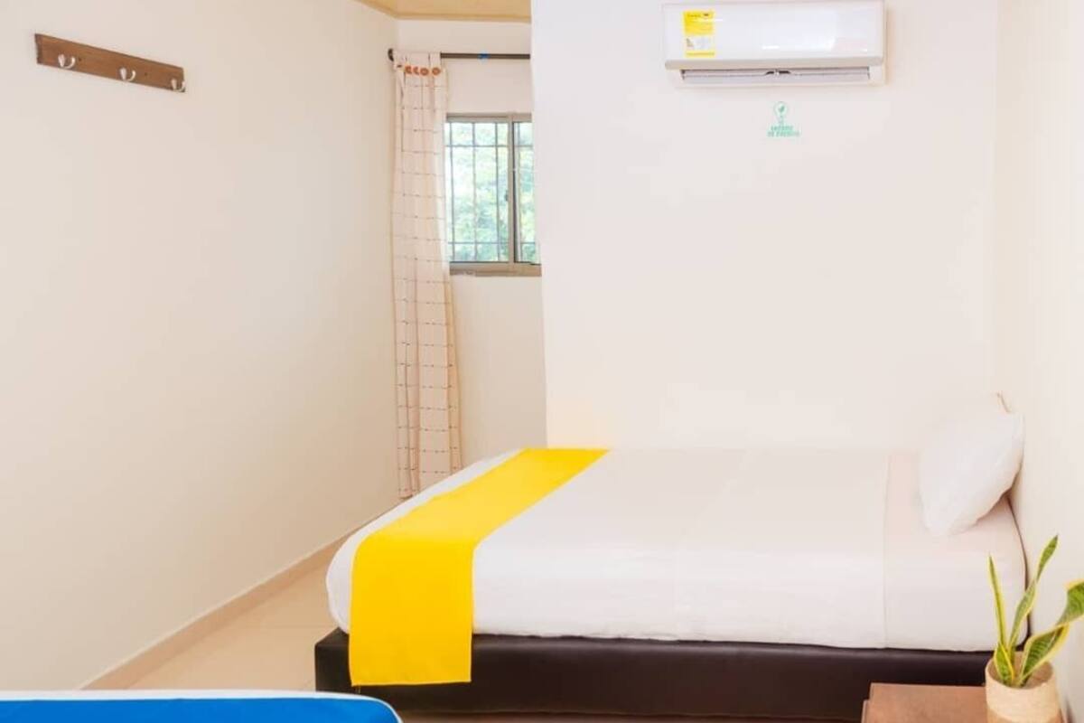A hotel room with a bed, a blue chair, a wooden nightstand with a plant, and an air conditioning unit on the wall.