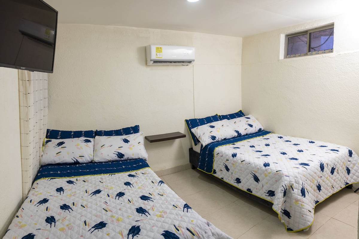 Two single beds with matching blue and white floral bedding, a wall-mounted air conditioner, and a small window with a view of the outside.